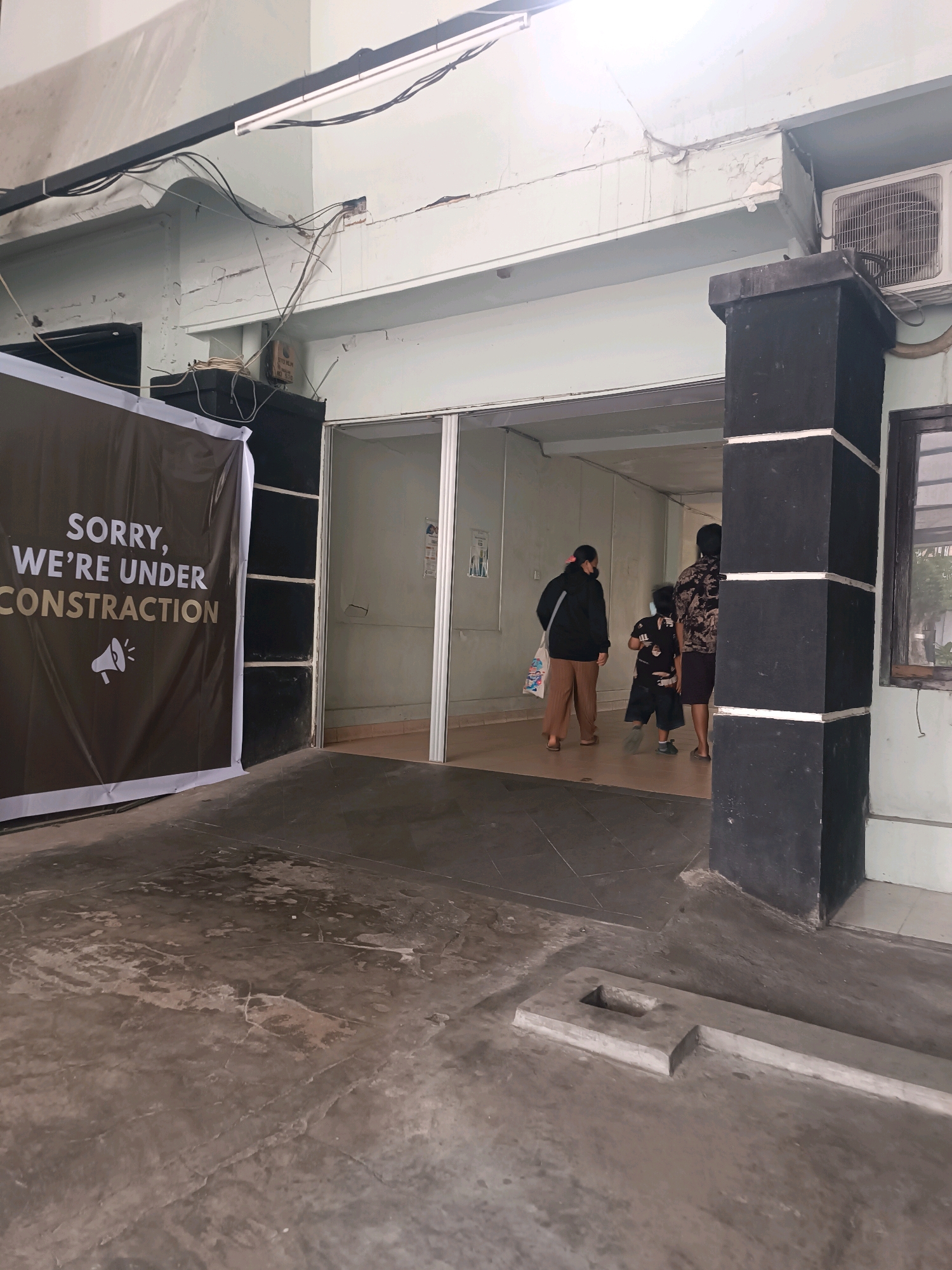 The image shows the exterior of a building with a sign that reads "SORRY, WE'RE UNDER CONSTRUCTION". The building appears to be located in Denpasar, Indonesia, based on the style of the architecture and the presence of an air conditioning unit commonly seen in tropical regions.
In the foreground, the ground is paved with concrete, showing signs of wear and tear with cracks and stains. To the left, a large dark brown banner with white and gold text is displayed, announcing the construction.
Further into the scene, an open doorway leads into the building, revealing a hallway. A black pillar with white stripes stands to the right of the entrance. Inside the hallway, three people are visible, walking away from the camera. The person furthest to the left is wearing a black jacket and brown pants, carrying a white bag. The middle person is a child wearing a black shirt with a pattern and dark shorts. The person on the right is wearing a patterned shirt and dark shorts.
The building's exterior wall is light-colored and shows some peeling paint and cracks. Electrical wires are visible, strung across the top of the building and connected to various conduits. An outdoor air conditioning unit is mounted on the wall above the entrance. A window with wooden framing is also visible on the right side of the building. The overall impression is of a modest, possibly older, commercial or institutional building undergoing renovation.