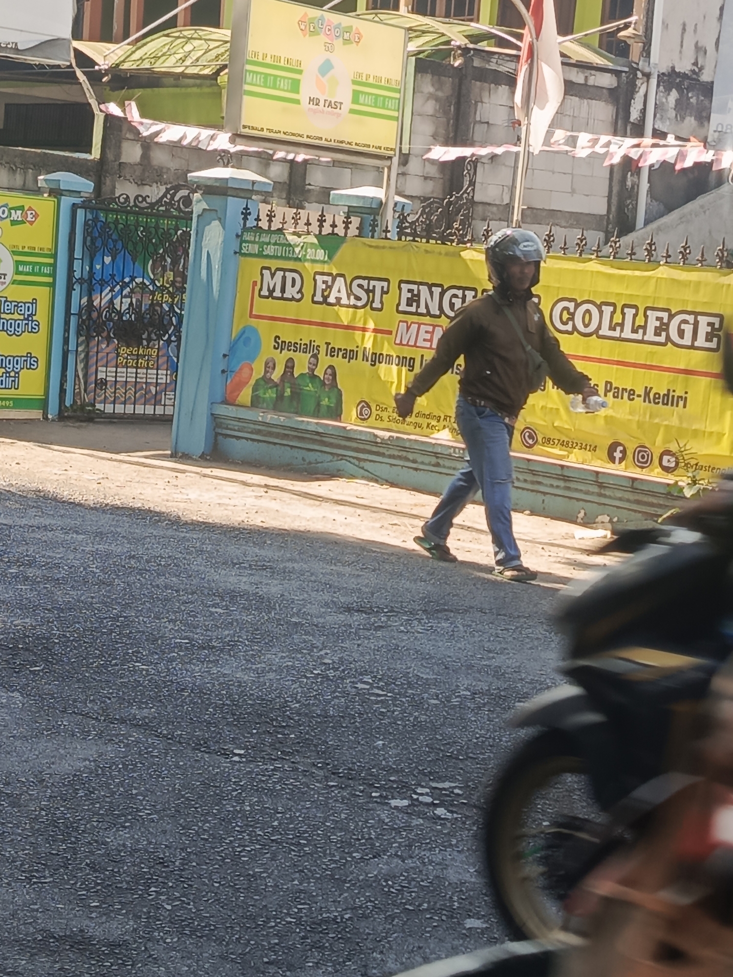 The image shows a street scene in Sidowungu, Indonesia, with a person walking across the frame. The person is wearing a dark jacket, blue jeans, and flip-flops, and is carrying a water bottle. They are walking in front of a banner that reads "MR FAST ENGLISH COLLEGE". To the left of the banner is a blue fence with a sign that says "Terapi Nggriss" and "Speaking Practice". The background shows a building with a green roof and a red and white flag. The street is paved with asphalt, and there is a motorcycle in the foreground on the right, partially obstructing the view. The image is taken in daylight, and the lighting suggests it is a sunny day.