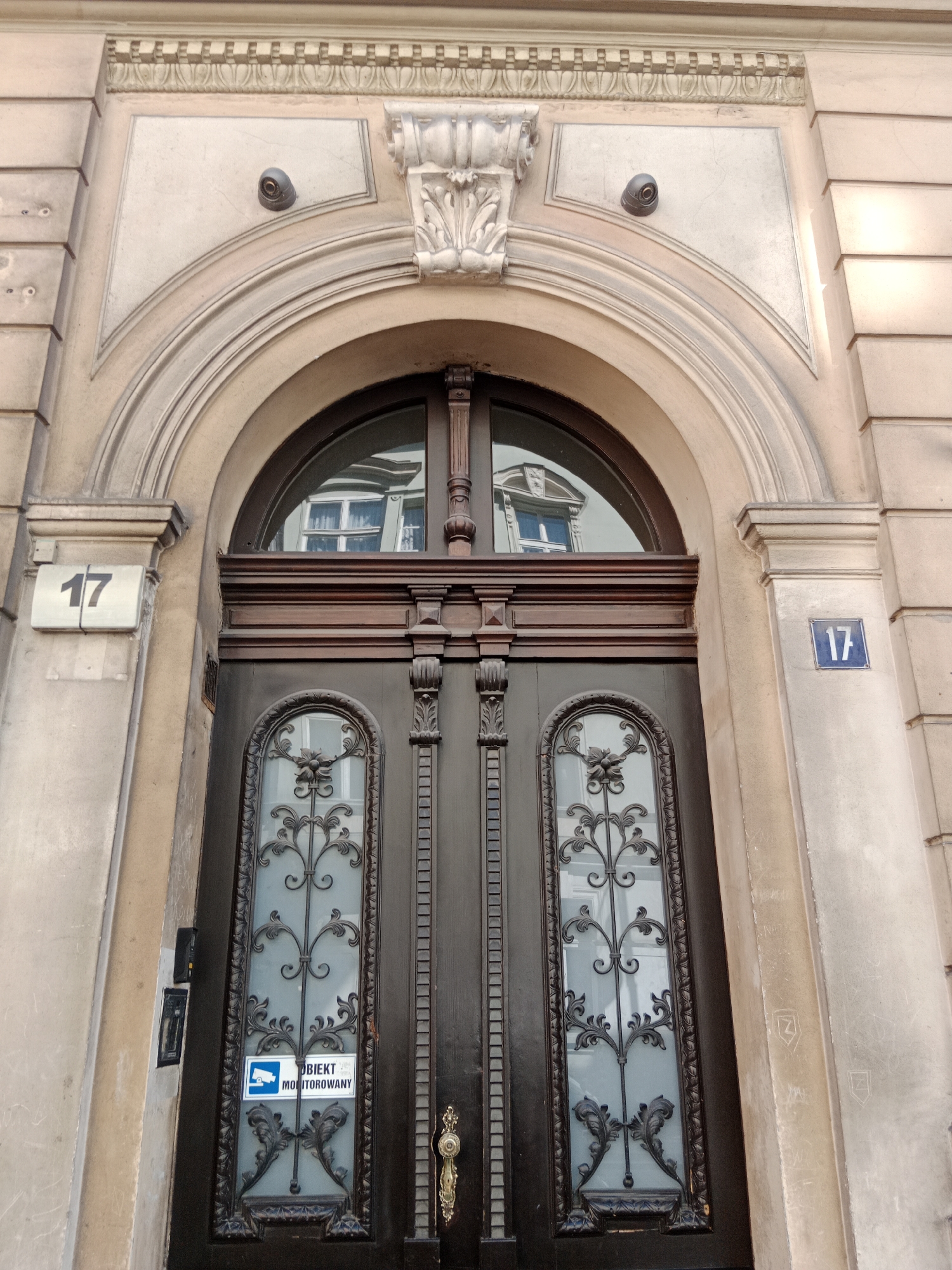 The image shows a dark wooden double door with ornate glass panels, set within a stone archway. Above the door is a semicircular window with reflections of buildings and windows. On the left and right sides of the archway, there are square blue and white address plaques displaying the number "17". On the upper part of the stone archway, above the door, are two small, dark, dome-shaped security cameras. Below the left camera, there is a decorative stone carving. On the left side of the door, attached to the stone pillar, is a small black intercom system. A blue and white sticker indicating "OBJECT MONITOROWANY" (monitored object) with a camera icon is affixed to the left glass panel of the door. The stone facade is a pale beige color, with some visible architectural detailing. There are no people visible in the image. This appears to be the entrance to a building at street number 17 in Bydgoszcz, Poland.