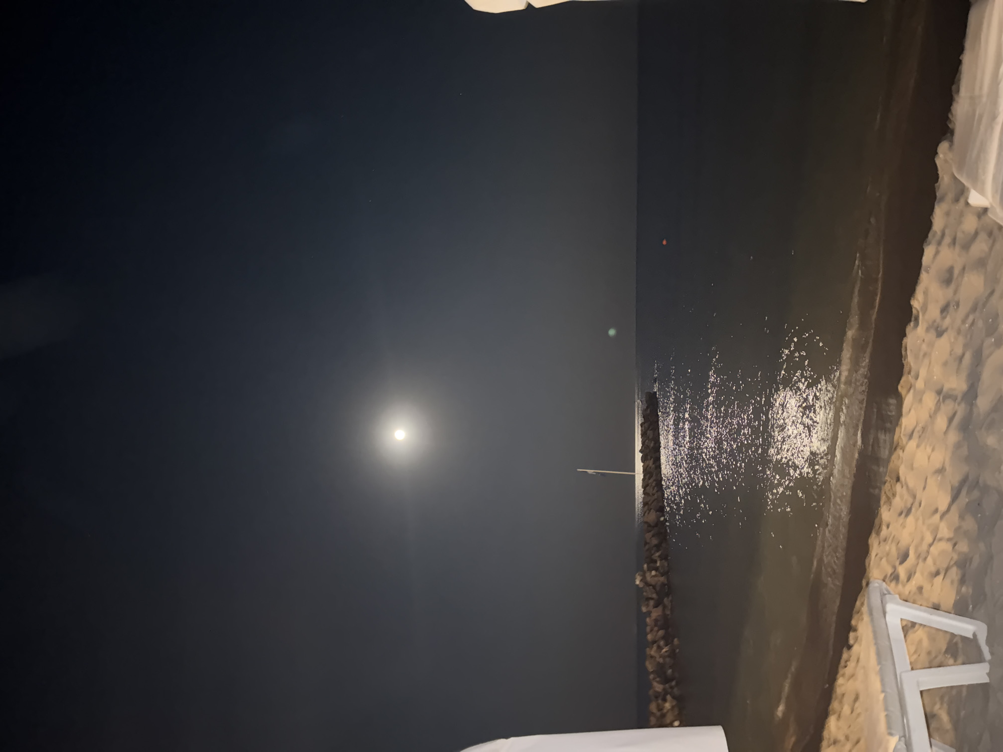 This is a night photograph of a beach scene. The sky is dark with a bright full moon casting a luminous reflection on the calm sea. In the foreground, a sandy beach is visible with a white lounge chair. A breakwater made of large stones stretches into the sea, and a flagpole stands on it. The water is dark and reflects the moonlight, creating a shimmering effect. The environment suggests a tranquil beach setting, likely in a coastal resort town. Based on the given information, this scene is located in Sveti Vlas, Bulgaria.