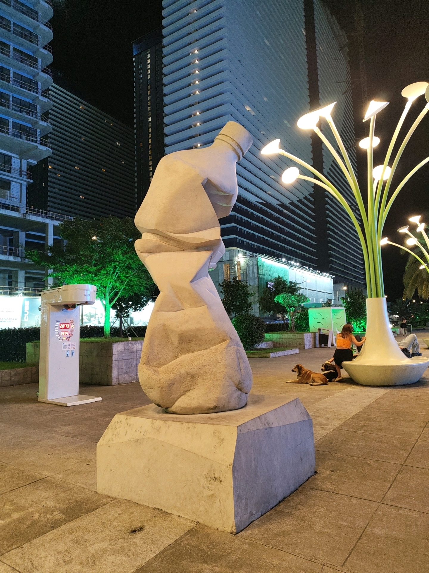 A large, abstract sculpture resembling a twisted, draped form sits on a concrete pedestal in an urban plaza at night. In the background, a modern, illuminated building with horizontal lines stands tall. To the right of the sculpture, a street lamp with multiple petal-like lights extends upwards from a large, white vase-shaped base. A person in an orange top and dark shorts leans against this base, while two dogs, one light brown and the other dark brown, lie on the ground nearby. To the left of the sculpture, a white, rectangular kiosk with a digital display is visible, featuring red and blue graphics. The plaza floor is paved with square tiles. The scene suggests a public space in Batumi, Georgia, likely an outdoor area of a modern development or entertainment district.