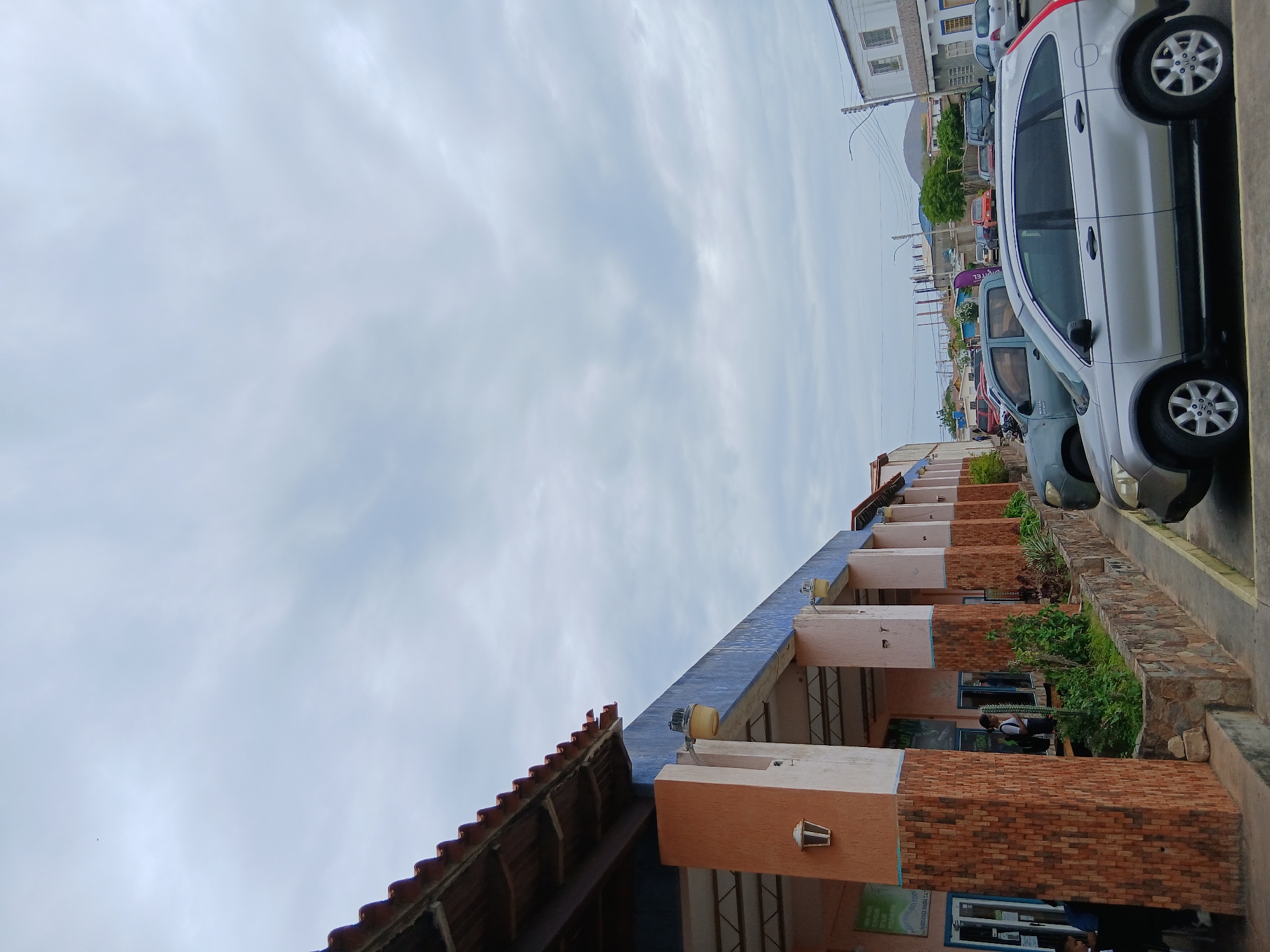 The image captures a street scene in Juan Griego, Venezuela, featuring a row of parked cars and a building with a tiled roof and brick columns. The sky is overcast. Several people are visible in the building's windows.