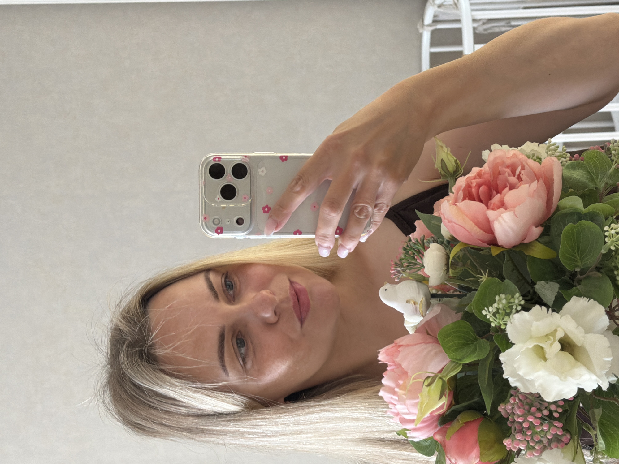 A blonde woman is taking a selfie in a room, holding a bouquet of pink and white flowers in her left arm. She is looking at her phone, which is also held in her left hand. Her right arm is slightly visible, positioned near the bouquet. The background is a plain, light-colored wall and a white crib. The image does not provide any information or visual cues to determine the location in Votkinsk, Russia.