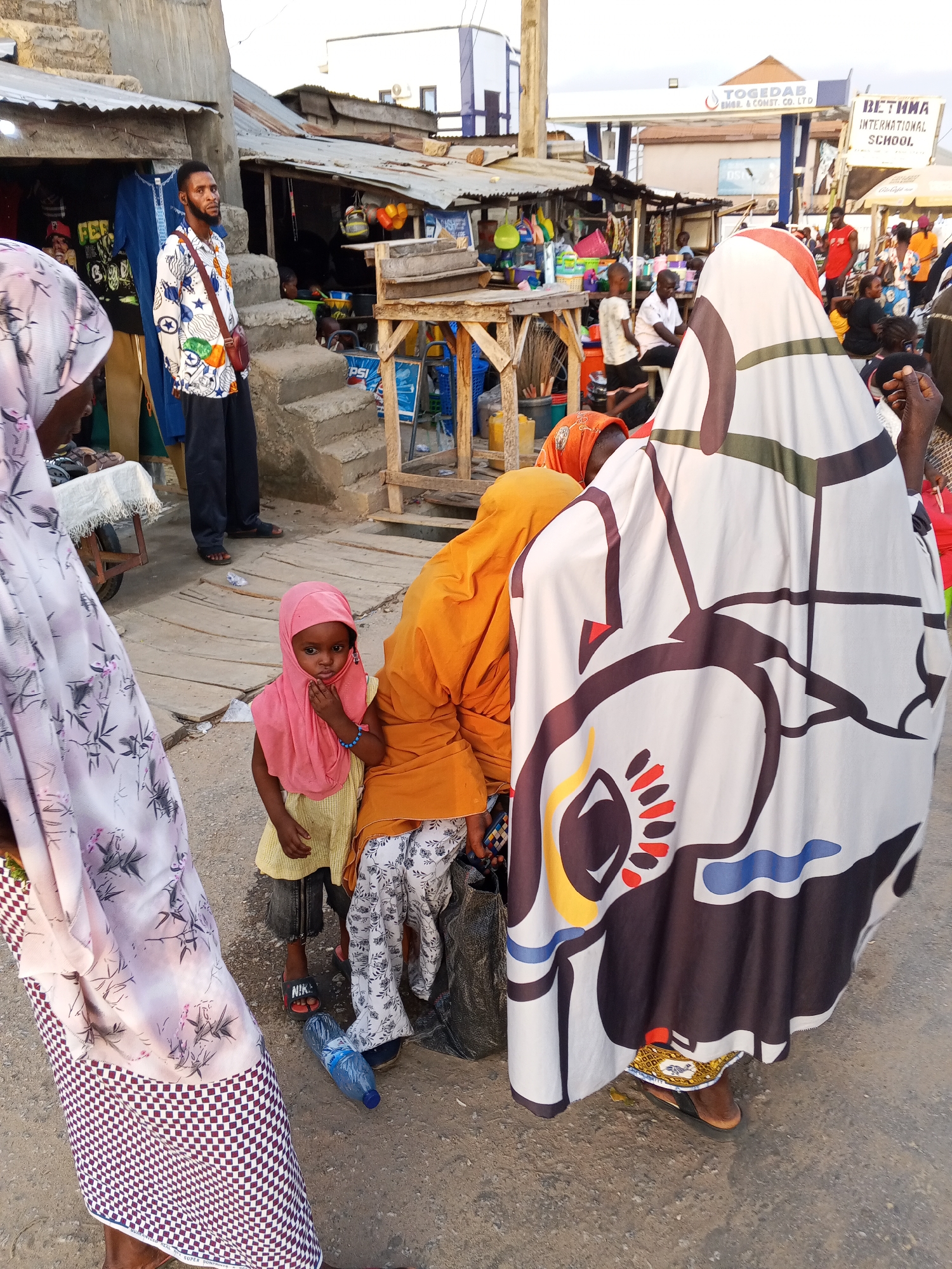 In the Zango-ndaji area of Nigeria, a group of people are gathered on a street. The focus is on a young girl wearing a pink headscarf and a yellow dress, who stands next to an adult woman in an orange garment. Another woman is dressed in a white robe with abstract designs. A plastic water bottle is on the ground nearby. In the background, there are market stalls, a man in a patterned shirt, and buildings, one of which has a sign for "Bethina International School". Other people are milling about, suggesting a busy, possibly commercial, environment.