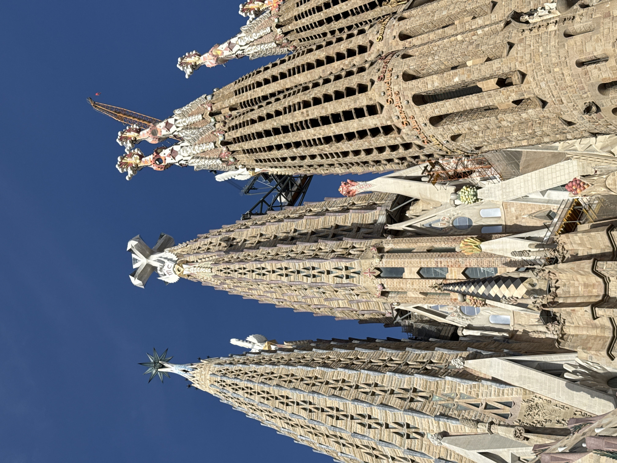 The image shows a low-angle view of the Sagrada Familia basilica in Barcelona, Spain, against a clear blue sky. The photograph focuses on the intricate spires and facades of the basilica, highlighting the detailed stonework and architectural ornamentation. Several spires are visible, each with unique designs and decorative elements, including crosses and star-shaped finials. Construction scaffolding and a crane are present, indicating ongoing work on the structure. The basilica's facade displays a rich texture of stone blocks and geometric patterns. Some inscriptions, possibly in Latin, are visible on the stone surfaces. The overall scene conveys a sense of grandeur and the ongoing process of completion for this iconic landmark.