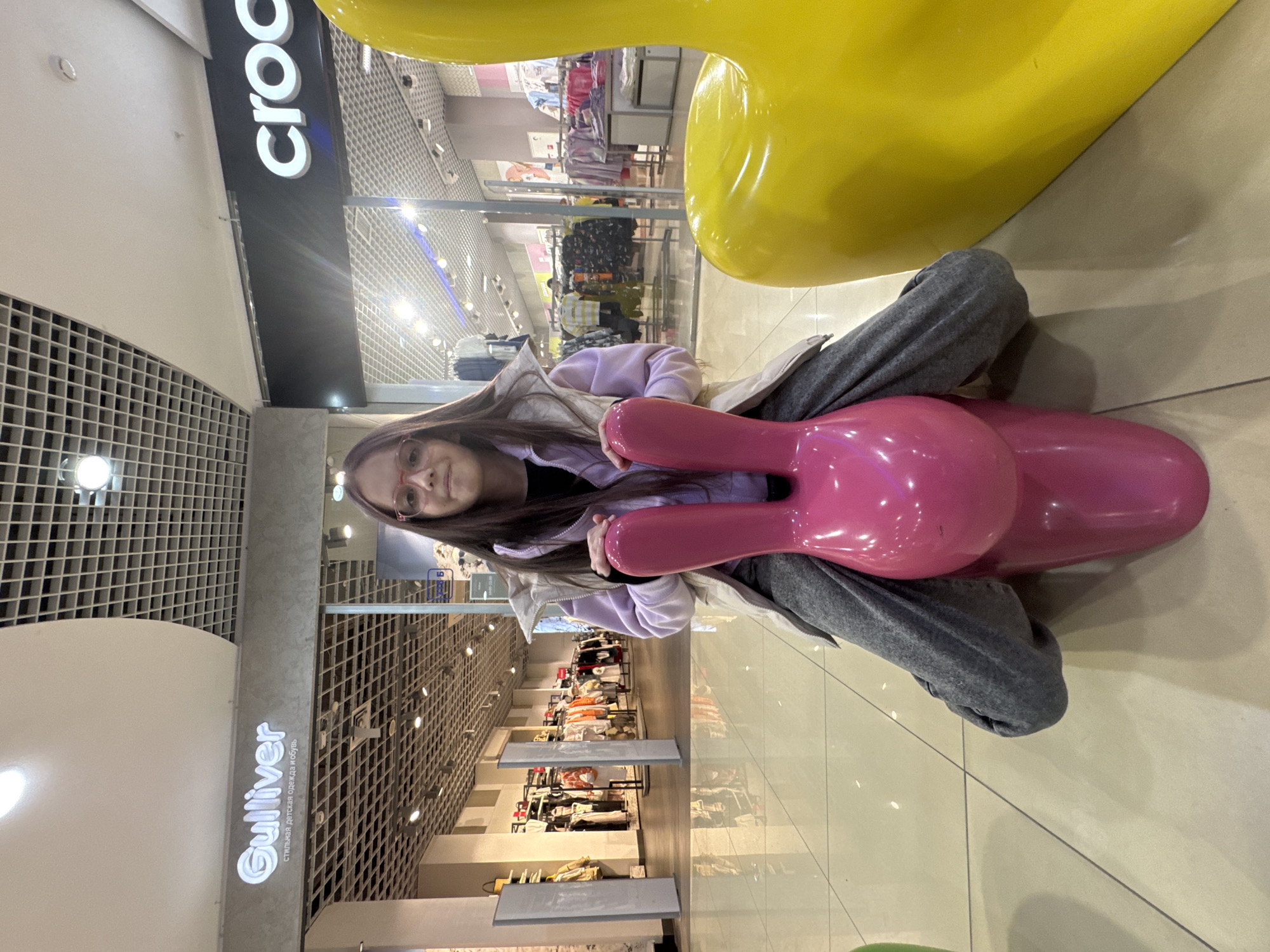 A girl is sitting on a pink, plastic rabbit seat. Beside the rabbit is a large yellow duck. The girl wears glasses, a black shirt, a light purple hoodie, and gray sweatpants. The location appears to be a shopping mall. Store signs visible are "Crocs" and "Gulliver".