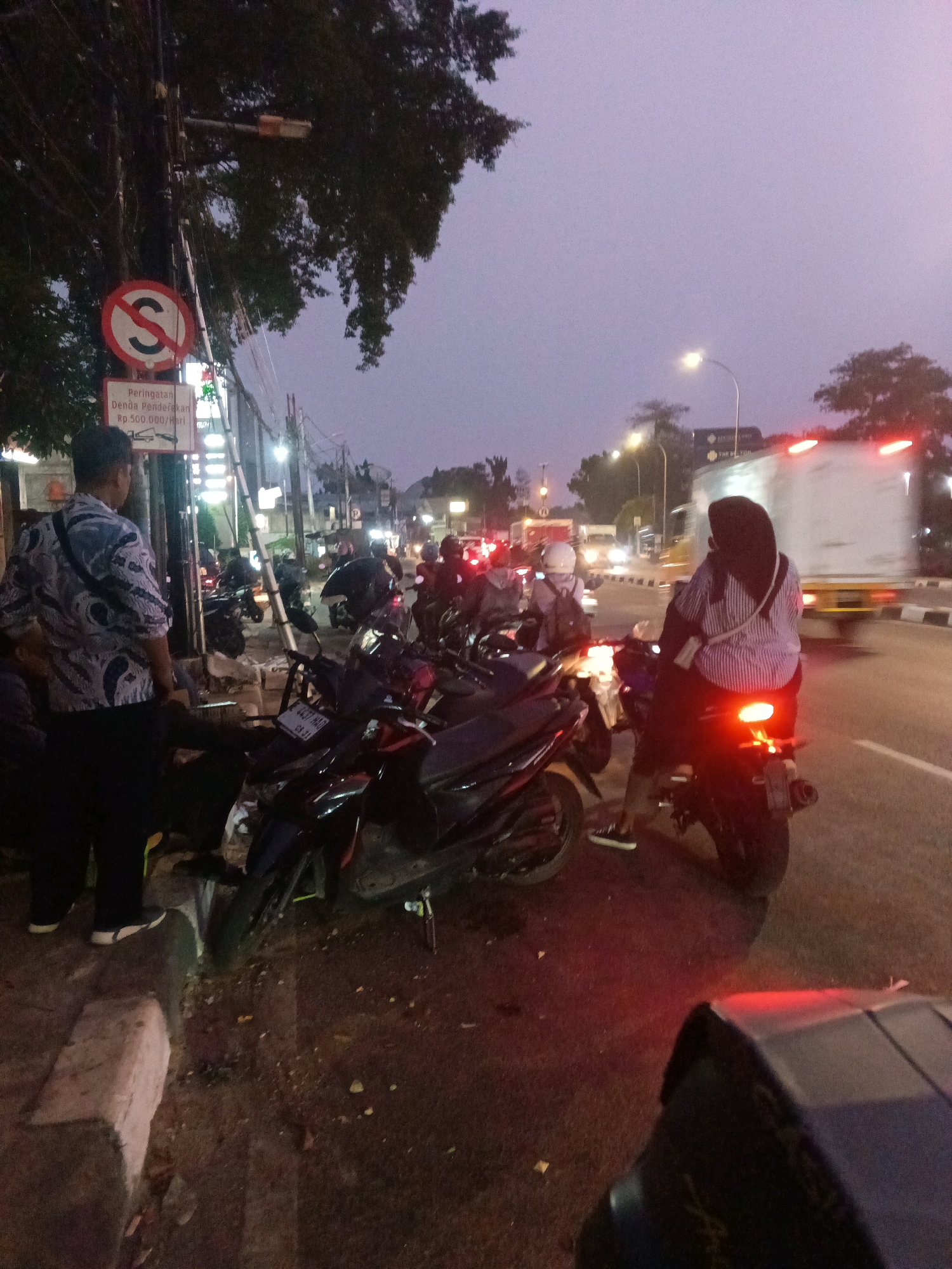 The image depicts a street scene in East Jakarta, Indonesia, during twilight or early evening. The foreground features a man in a patterned shirt and dark pants standing near a sidewalk, facing away from the camera. Several motorcycles are parked along the roadside, some with their lights on. In the background, a large white truck is visible on the road, along with other vehicles and streetlights. The sky is a dusky purple, and there is a traffic sign with a red circle and a diagonal line, indicating "No Stopping" or "No Parking". A sign below it reads "Peringatan Denda Parkir Rp.500.000/Hari", which translates to "Parking Fine Warning Rp.500,000/Day". The overall impression is of a busy urban environment with traffic and pedestrians.