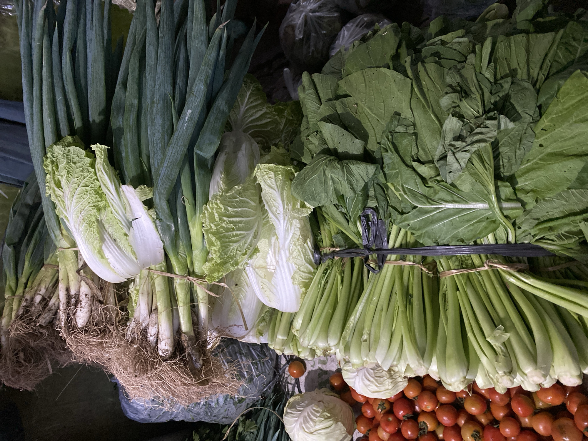 The image displays a fresh produce stall in Ambarketawang, Indonesia, featuring a variety of vegetables. Prominently displayed are bundles of green onions with their roots still attached, several heads of Napa cabbage, and bunches of green leafy vegetables, possibly bok choy or similar. A pile of small, red cherry tomatoes is visible on the left, alongside a round head of white cabbage. The vegetables are arranged in stacks, some secured with black ties. The background is dark and appears to be the interior of a market stall.