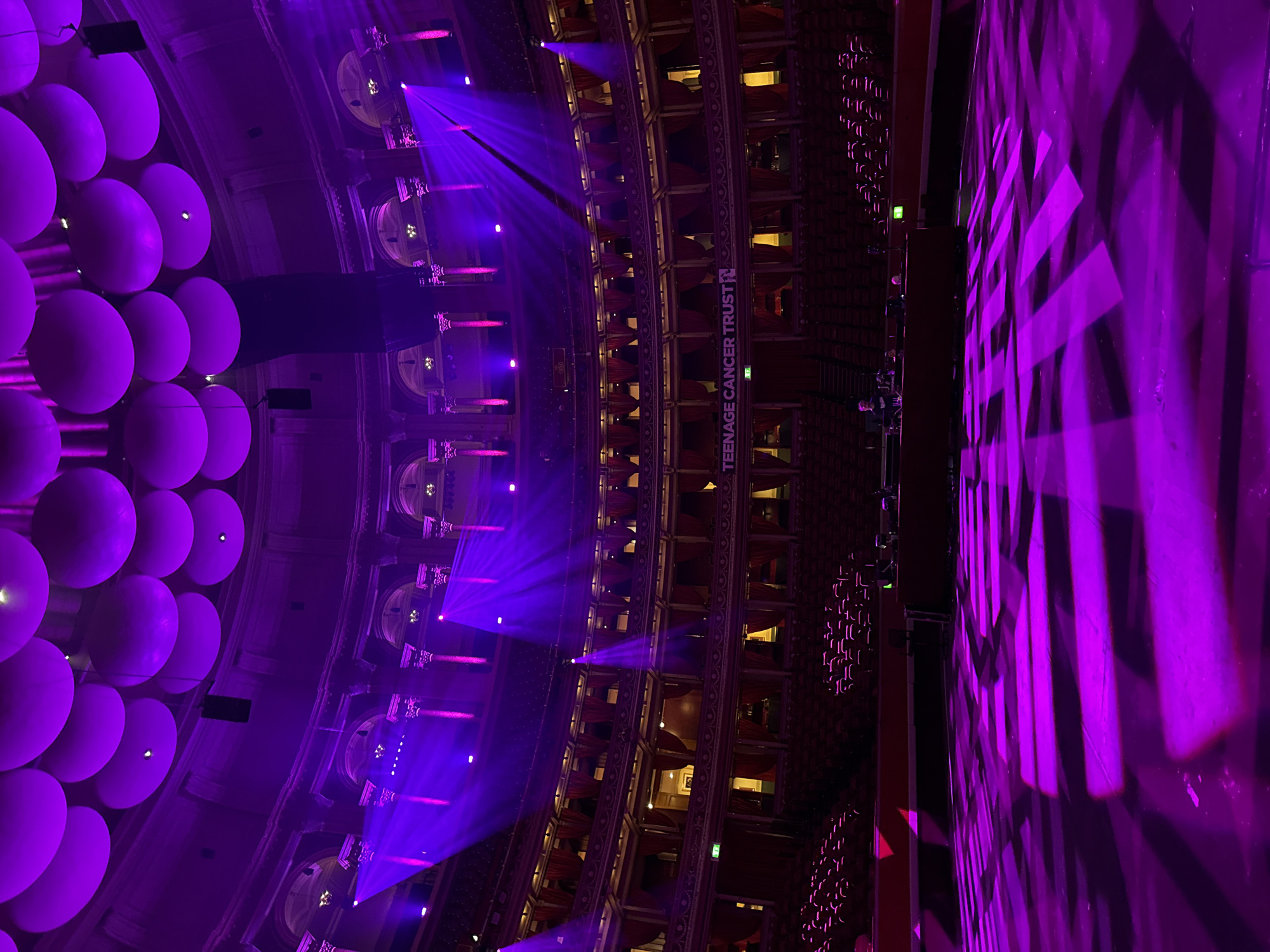 The image displays the interior of the Royal Albert Hall in London, United Kingdom, illuminated predominantly by purple stage lighting. The perspective is from the main stage, looking up towards the tiered, circular auditorium. The domed ceiling features numerous large, spherical or disc-shaped light fixtures or acoustic panels. Multiple levels of balconies, adorned with ornate columns and archways, encircle the hall, presenting rows of unoccupied red velvet seating and boxes with red drapes. A white banner with the text "TEENAGE CANCER TRUST" is prominently displayed on a middle balcony. On the stage floor in the foreground, geometric patterns of purple light are projected. In the mid-ground on the stage, a dark, rectangular console or barrier is visible, behind which two individuals are partially observed interacting with equipment. Beams of intense purple light emanate from various stage lighting fixtures, projecting across the empty auditorium.