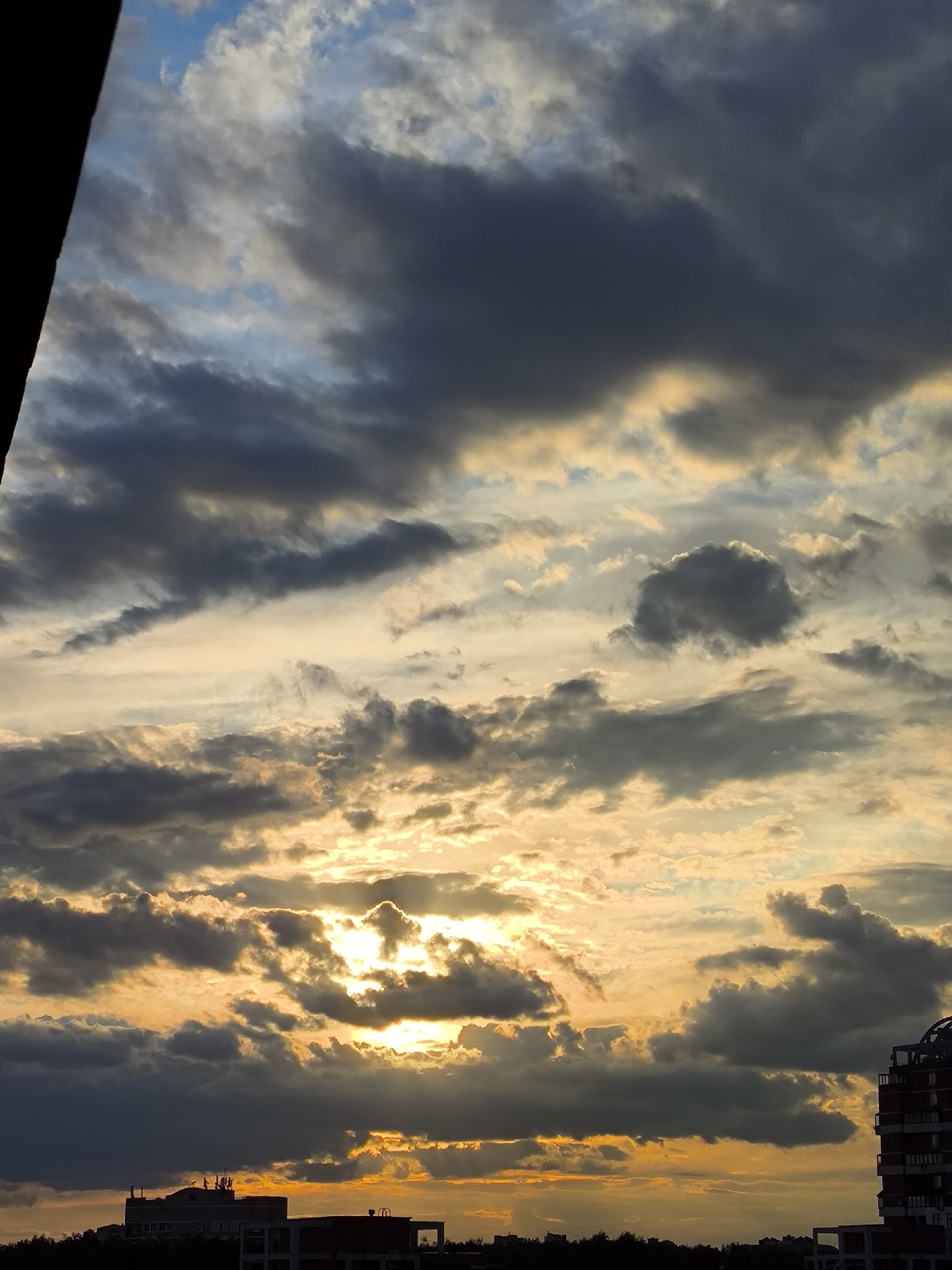 The image presents a dramatic and colorful sky during what appears to be either a sunrise or sunset, casting a warm glow over a silhouetted urban or suburban landscape. The sky is a prominent feature, filled with a complex arrangement of clouds. Patches of clear blue sky are visible in the upper left, but the majority of the sky is covered by thick clouds ranging in color from dark grey and blue-grey to bright white and vivid gold.
The sun is partially obscured by these clouds in the lower-middle section of the sky, creating a powerful burst of golden light that illuminates the surrounding cloud edges and projects visible sun rays downwards. The golden and orange light extends across the middle and lower parts of the sky, contrasting sharply with the darker, heavier cloud masses above.
At the bottom of the frame, the skyline of buildings and trees is rendered in a dark silhouette against the brightly lit sky. On the left, a multi-story building with apparent rooftop structures or antennae is visible. On the right, another building with a more distinct architectural form, possibly featuring a rounded element on its roof and visible balconies, stands out. Additional smaller structures and the outlines of trees are also discernible along the horizon. A dark, angular object intrudes into the top-left corner of the image, suggesting it might be part of a window frame or another foreground element. The scene evokes a sense of awe and natural beauty during a transitional period of the day. No text is visible in the image.