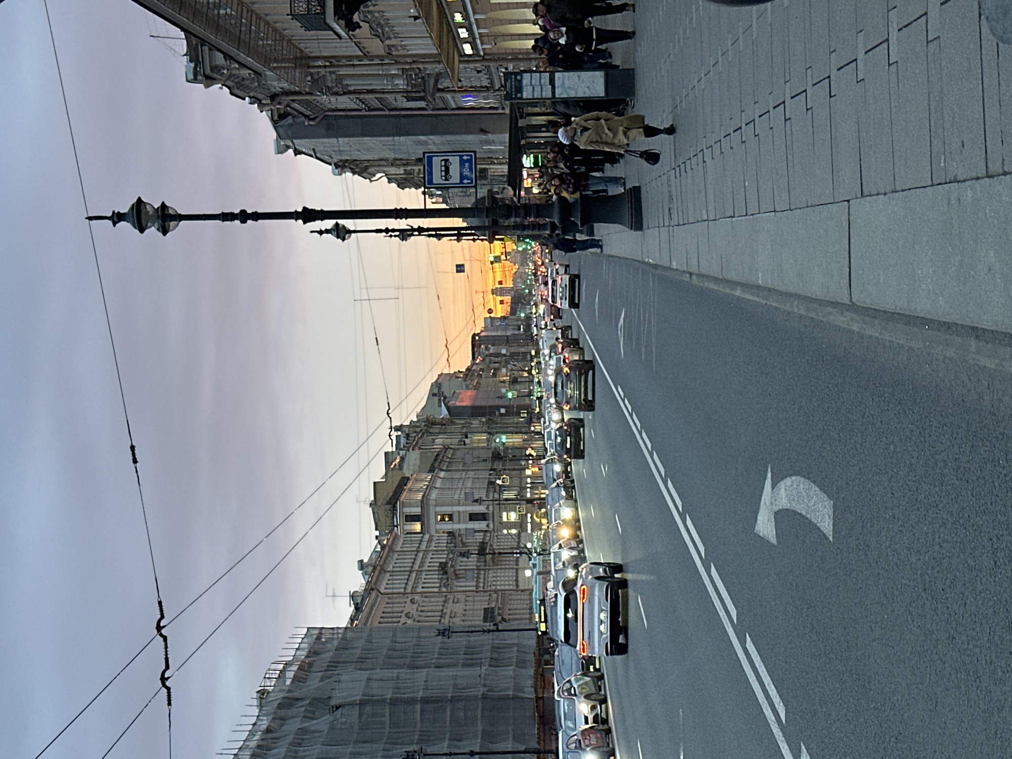 The image shows a street scene in Saint Petersburg, Russia, at dusk or dawn, with a line of cars in a traffic jam extending into the distance. Buildings line the sides of the street, some of which are undergoing construction and are covered in netting. Pedestrians stand and wait on the sidewalk. Streetlights line the road. The street has white painted directional markings.