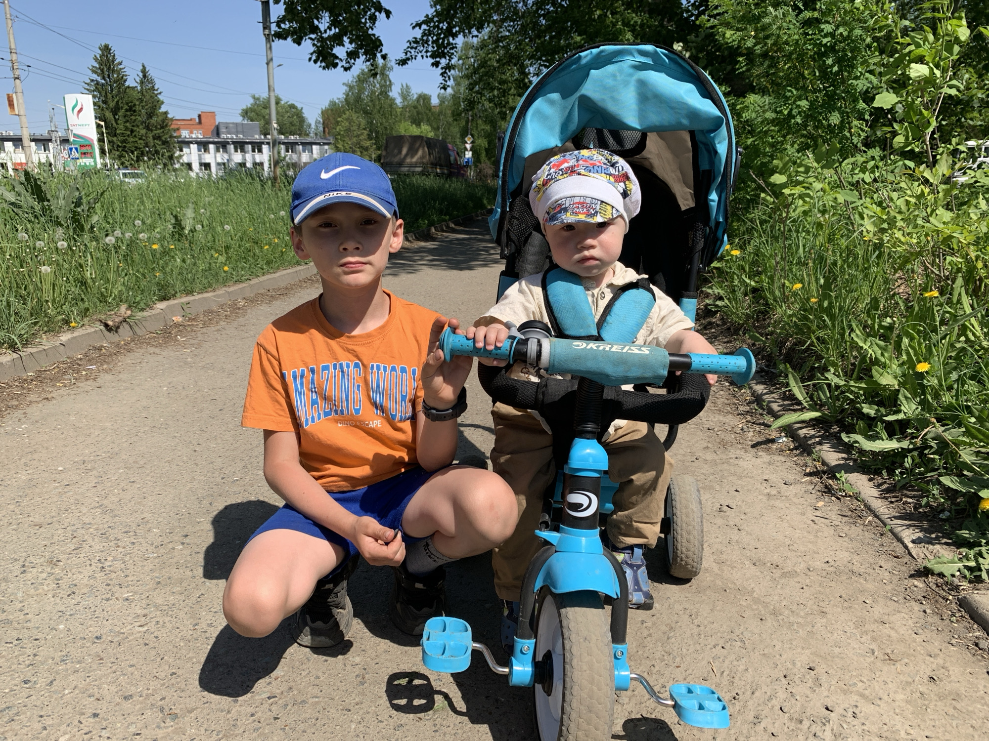Two boys are on a path in Izhevsk, Russia. One boy, wearing a blue Nike baseball cap, an orange t-shirt, and blue shorts, is crouching on the path. The other boy is sitting on a blue tricycle with a light blue sunshade. He is wearing a patterned cap and a light tan shirt. The tricycle has the brand name "OKREISS" on the handlebar stem. The path is paved with asphalt and bordered by grass and dandelions on one side and bushes on the other. In the background, a building with a green and white sign that reads "TATNEFT" is visible.