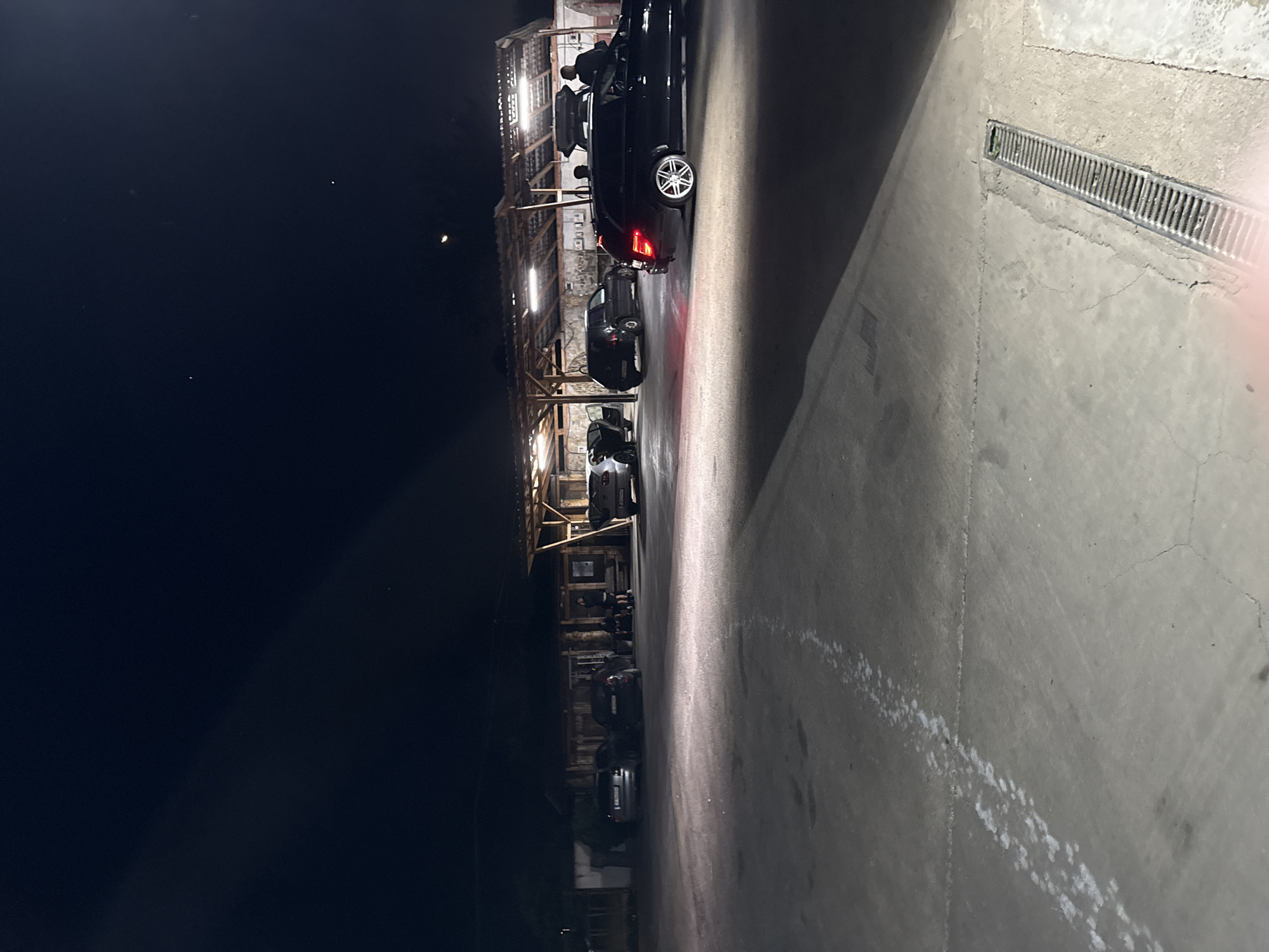 The image shows an alleyway at night in Elhovo, Bulgaria. Several dark-colored vehicles are parked under a wooden structure. At least two people are visible near the first vehicle, with its doors open. The ground is concrete and shows tire tracks and dirt. A metal drain is visible on the right side. The sky is dark with few visible stars. The lighting is predominantly artificial, coming from the structure above the vehicles.