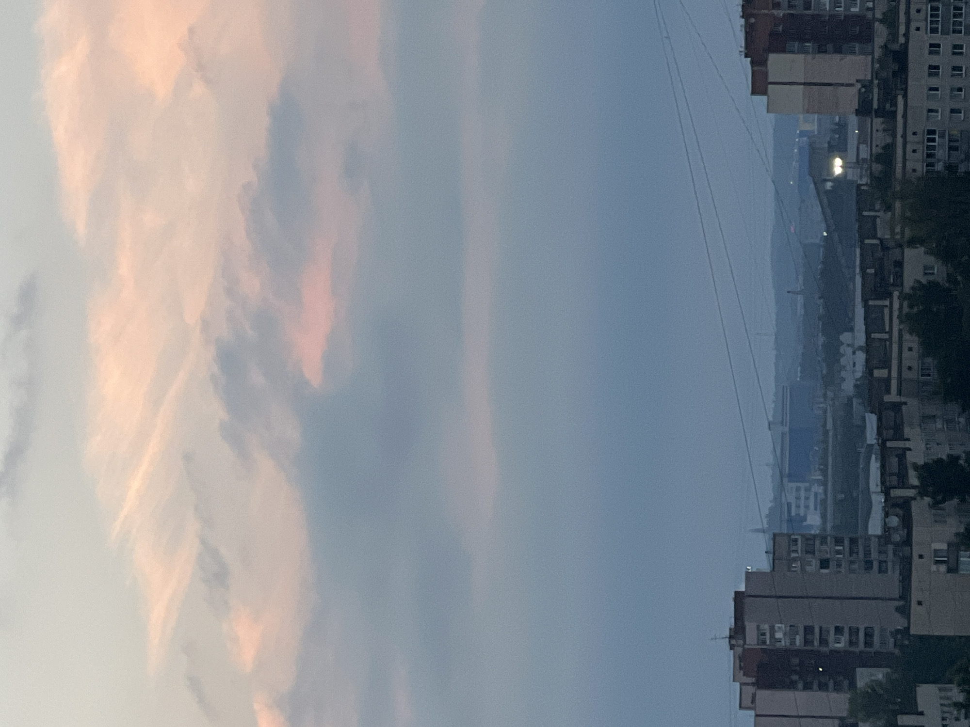 An aerial perspective of a city skyline at dusk. The sky is filled with streaky clouds tinged with pink and orange from the setting sun, transitioning to a muted blue towards the horizon. In the foreground, apartment buildings with visible windows and balconies line the cityscape. Beyond these, a hazy industrial or commercial area with larger, boxy structures is discernible. Power lines crisscross the upper portion of the image. The scene depicts an urban environment under a twilight sky.