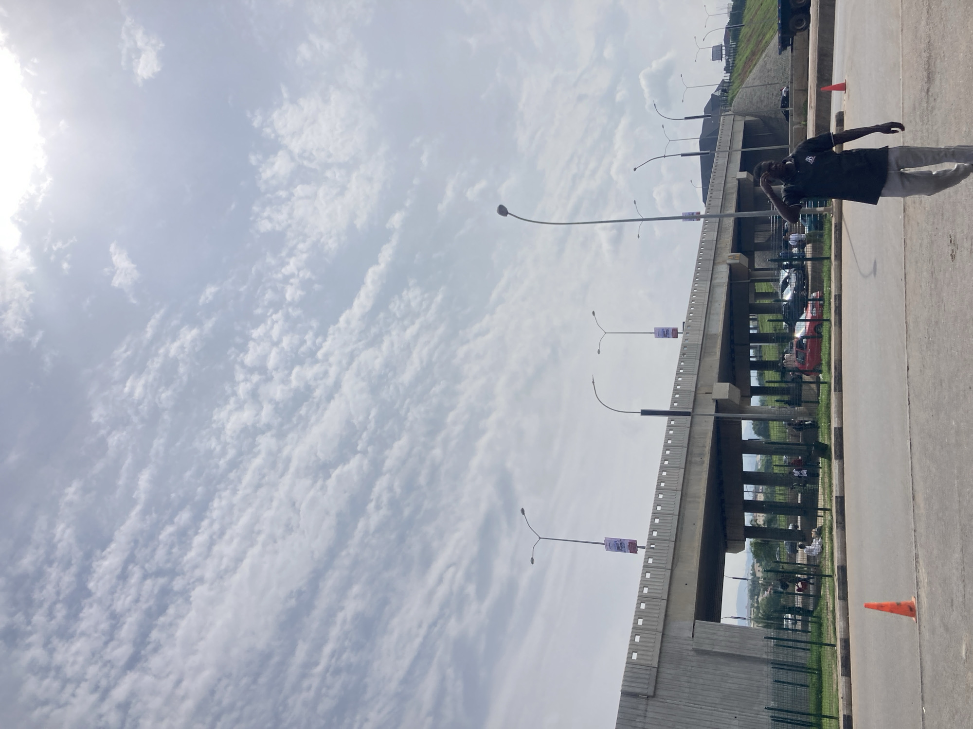 The image captures an outdoor scene dominated by a concrete overpass or bridge under a bright, partly cloudy sky. The sun is visible in the upper left, casting a bright light across the scene, suggesting daytime.
In the foreground, a paved road stretches across the bottom of the frame, with an orange traffic cone visible on the right side. On this road, a man stands on the right, clad in a dark short-sleeved collared shirt and light-colored trousers. He appears to be looking towards the left, with one hand raised towards his face or head, in a contemplative or perhaps pensive posture.
The prominent overpass structure runs horizontally across the mid-ground, supported by pillars. Street light poles with curved arms and light fixtures extend upwards from the overpass, and similar lights are also visible along the road in the foreground. Small rectangular banners or signs are attached to some of these poles on the overpass, but their text is illegible due to distance and resolution.
Beneath the overpass, there's a visible area with parked cars and several people standing or sitting on what appears to be grass or a lower ground level. A wire mesh fence separates the foreground road from the area under the bridge. To the left of the overpass, a green, grassy embankment slopes upwards, with a large truck partially visible at its crest.
The sky above is filled with scattered, patchy clouds, indicative of a bright but not entirely clear day. The general setting, with the large infrastructure and varied activities, suggests an urban or peri-urban environment, consistent with the context of Abuja, Nigeria.