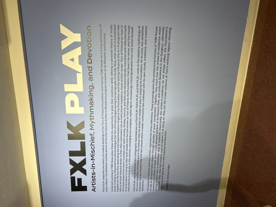 A poster advertises the "FXLK PLAY" exhibit, which celebrates the history of creative exchange at the Tom of Finland Foundation. The exhibit reunites alumni of the international Artists-in-Residence program in honor of its fifteenth anniversary, exploring the erotic power and cultural dialogue begun by Tom of Finland. The exhibition features sixty-four artists and the architectural fixtures of TOM House. The exhibit explores themes of mythmaking, mischief, and devotion and is curated by Jamison Edgar.