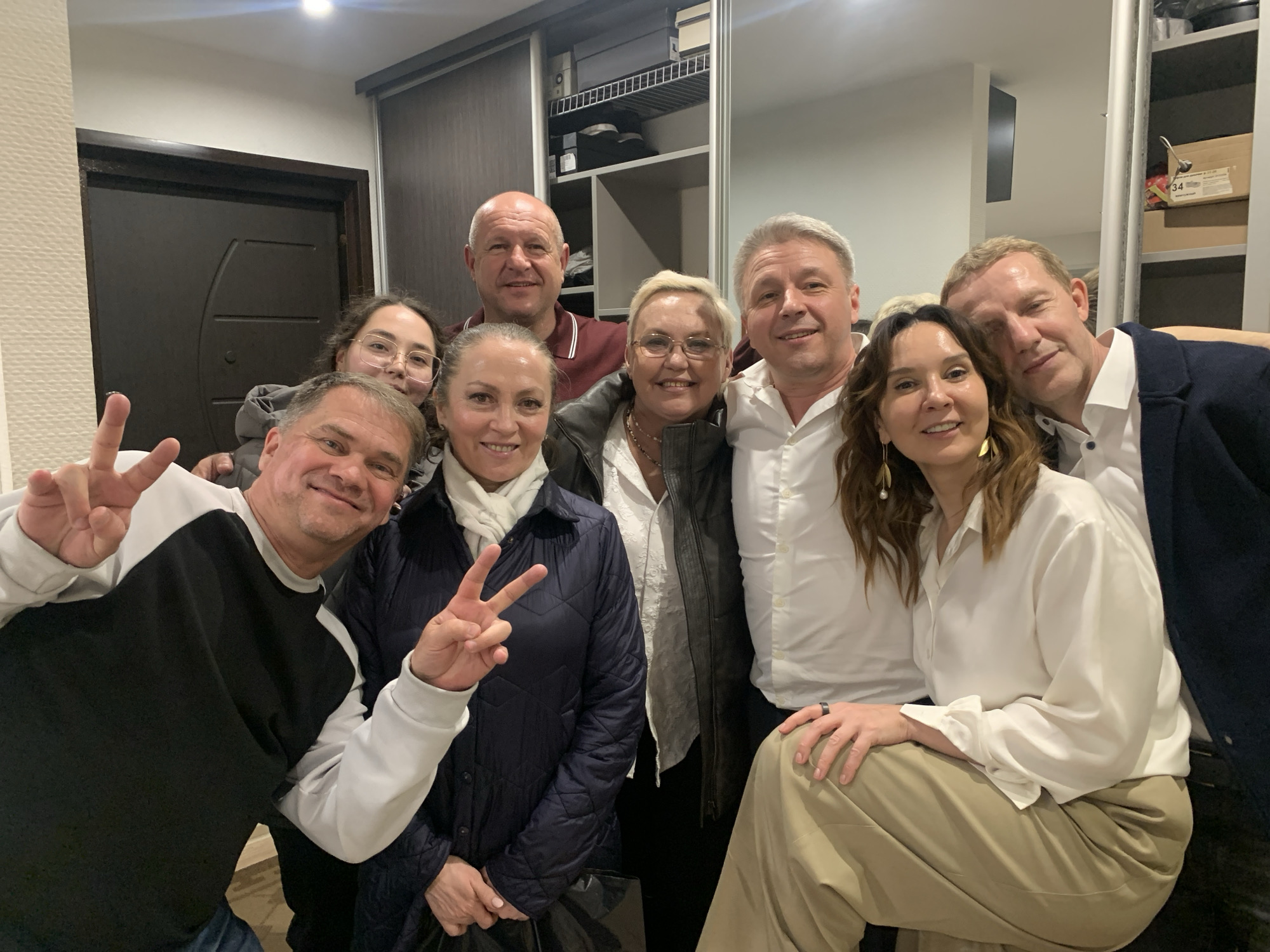 A group of eight people pose closely together in an indoor setting, likely a private residence. The group comprises a mix of men and women of varying ages. One man in the foreground is making a peace sign with his fingers. The background includes a door, a wardrobe with mirrored panels, and shelving with storage boxes. The location is Chaikovsky, Russia.