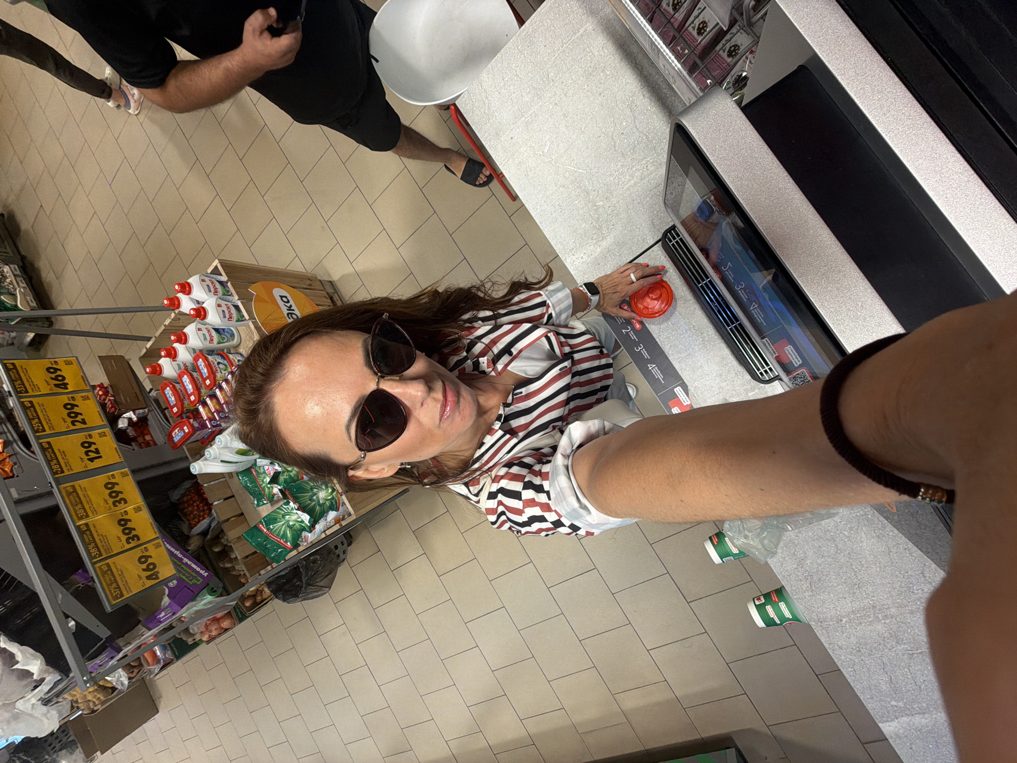 A woman is taking a selfie in a supermarket in Birobidzhan, Russia. She is wearing a striped shirt and sunglasses. In the background, there are shelves stocked with various products, including cleaning supplies and produce. A man in black shorts and a black t-shirt can be seen in the background. The woman is holding a red cup.