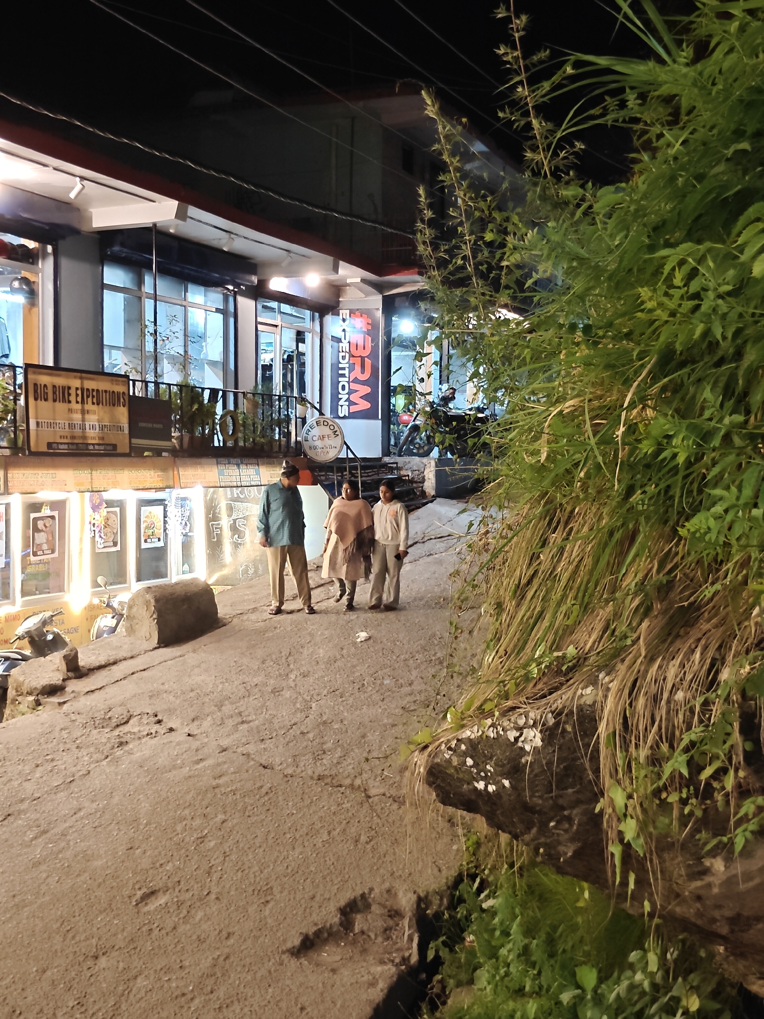 This is a nighttime photograph of a street scene in Vashisht, India. Three people are walking up a sloped, paved path in front of several businesses. The person on the left wears a teal shirt and tan pants, the middle person wears a light pink shawl over a white outfit, and the person on the right wears a light tan hoodie and pants.
The businesses have large glass windows and signs. One sign reads "BIG BIKE EXPEDITIONS MOTORCYCLE RENTALS AND EXPEDITIONS." Another sign, vertically oriented and partially obscured by vegetation, reads "#BRM EXPEDITIONS." A smaller sign advertises "FREEDOM CAFE 8:00 AM - 11 PM OPEN." The area appears to be a commercial district with shops and a cafe, illuminated by artificial lights, suggesting it is evening or night.
To the right of the path, a dense area of tall, dry grass and green foliage grows on a rocky incline, partially obscuring the background buildings. There are rocks and debris on the ground, indicating an uneven terrain. A motorcycle is parked near the businesses on the left. The overall atmosphere is casual and everyday, with the activity being pedestrian movement through the illuminated street.