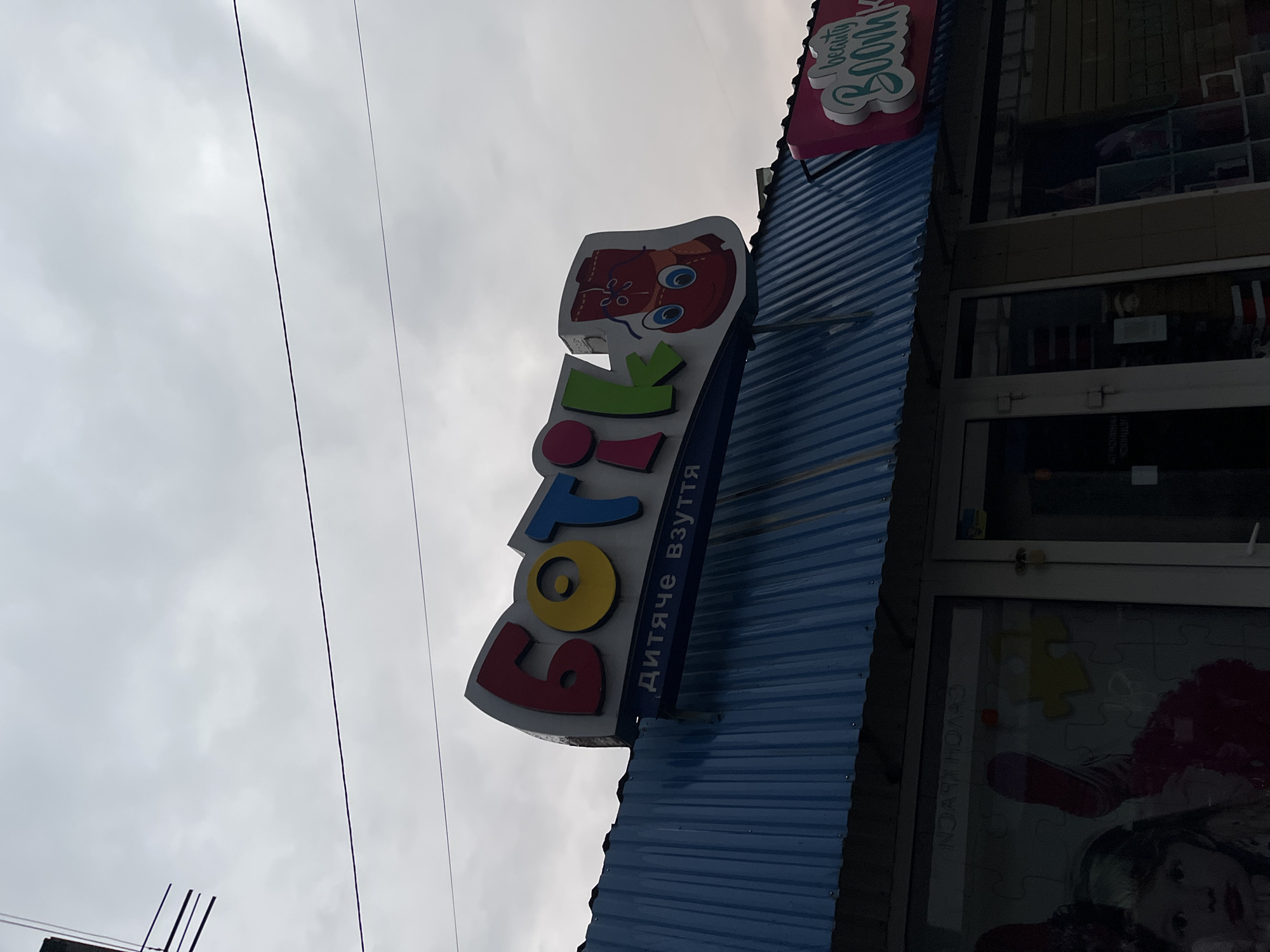 This is a medium-angle, eye-level, full exterior shot of a storefront in Chernivtsi, Ukraine. The sky is overcast with a muted gray appearance. In the foreground, a blue corrugated metal awning covers the shop. Above the awning, a large, colorful sign for "БОТІК" is prominently displayed. The letters are three-dimensional and brightly colored: "Б" is red, "О" is yellow, "Т" is blue, and "ІК" are green. To the right of the letters, a cartoon illustration of a red boot with a smiling face is part of the sign. Below the main sign, a smaller sign reads "ДИТЯЧЕ ВЗУТТЯ" in white letters. To the right of the "БОТІК" sign, another smaller pink sign with white lettering says "beauty BOOM". The storefront windows on the left are dark and reflective, showing the interior of the shop. A young girl's face is visible on a poster in the left window. The shop entrance is visible in the center, with a white door and glass. The overall impression is that of a children's shoe store.