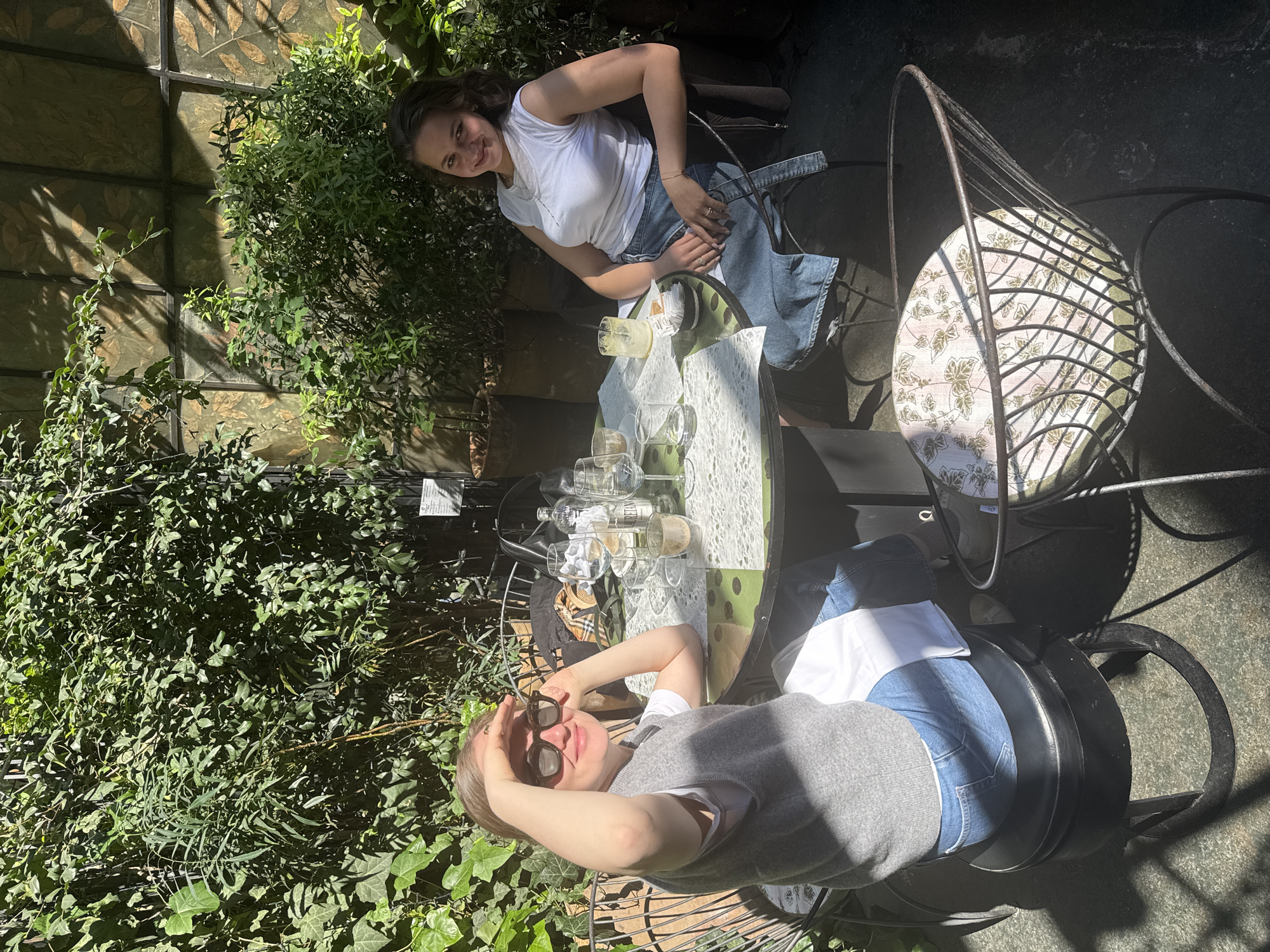 This image depicts two women enjoying drinks at an outdoor patio in Milan, Italy, on a bright, sunny day. The scene is lush with green foliage, including dense bushes and climbing ivy, which provides a natural, enclosed setting. Strong sunlight casts clear shadows, indicating mid-day or afternoon.
On the left side of the round, light green table, a woman with dark hair is seated, smiling broadly at the camera. She wears a white t-shirt and a denim-style apron or skirt, and is seated in a metal chair with a light-colored, patterned cushion.
Opposite her, on the right, another woman with lighter hair is seen with sunglasses on, casually shielding her eyes from the sun with her hand while smiling. She is dressed in a grey vest over a white t-shirt and blue jeans, sitting in a black metal chair.
The table between them holds several items, including multiple wine glasses (some empty, others containing white wine or a similar beverage), and a tall glass with an iced yellow drink and a lemon wedge. White paper napkins or coasters are also visible. Both women appear relaxed and happy, suggesting a pleasant social gathering. Some small text is visible on a card on the table and possibly a sign in the background, but it is illegible.