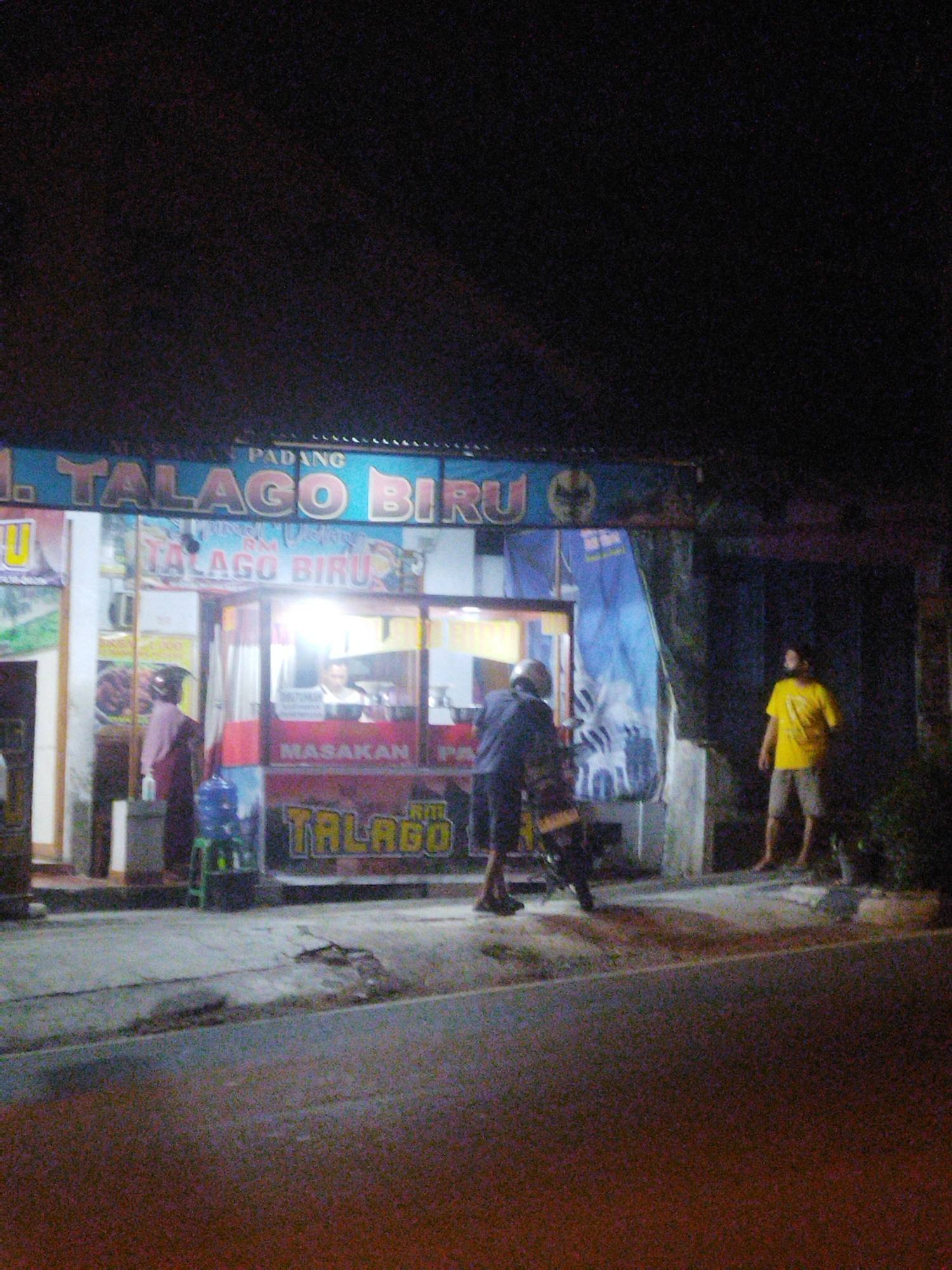 The image depicts the exterior of a food stall or small restaurant at night. A neon sign above the establishment reads "PADANG" and "TALAGO BIRU". The building has a transparent front with a counter, where a person appears to be working. Outside the building, to the left, a person in a maroon outfit stands near a blue water dispenser. To the right, a person wearing a yellow t-shirt and shorts stands on the sidewalk. A motorcycle is parked in front of the stall, with another person leaning over it. The ground is paved with a rough texture, and there are some damaged sections. The overall lighting suggests it is nighttime, with artificial lights from the stall illuminating the immediate area. The scene is likely in Wonogiri, Indonesia, as indicated by the signage.