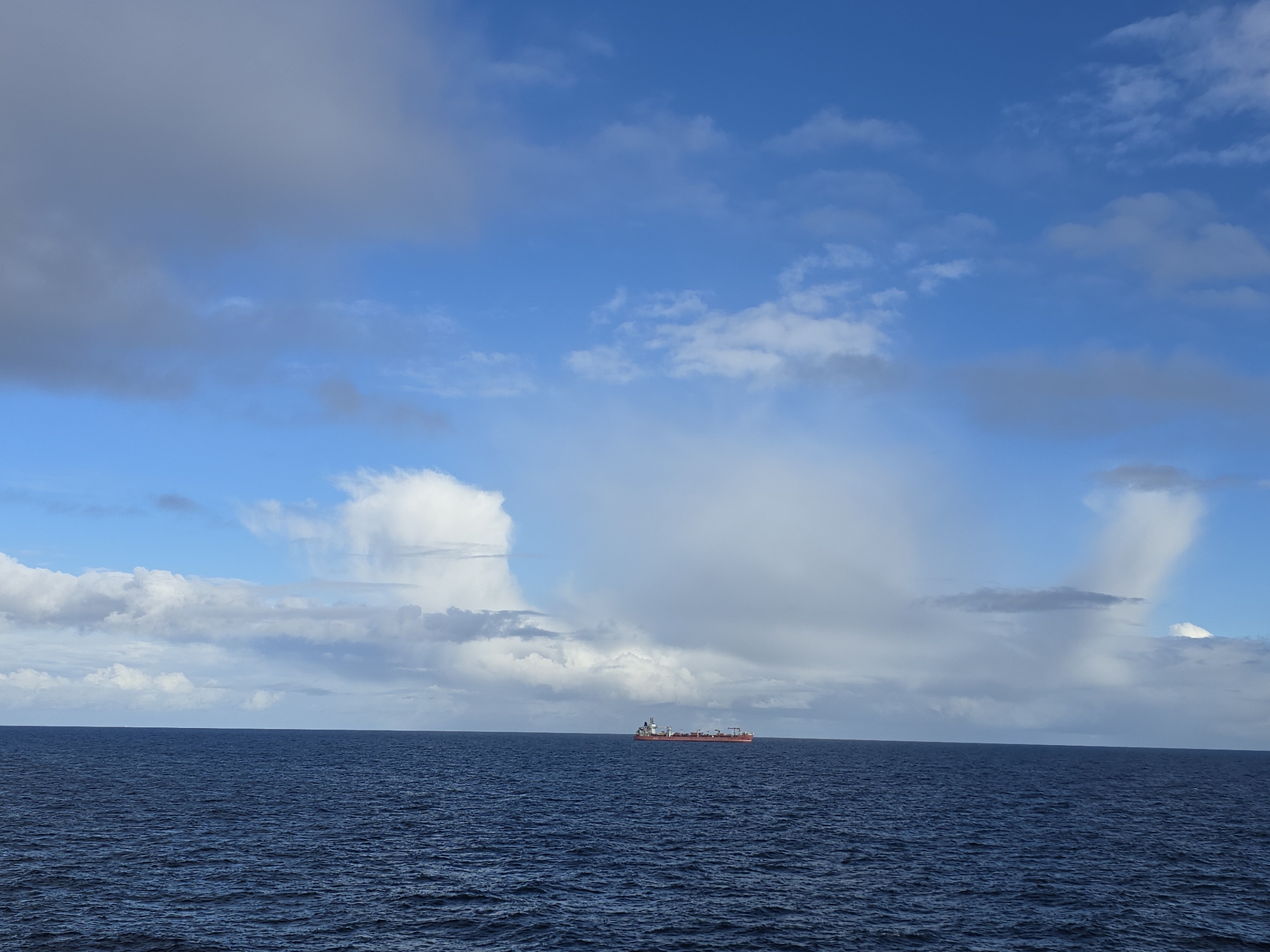A ship floats on the ocean under a blue sky with various cloud formations. Location: Unknown, Unknown.