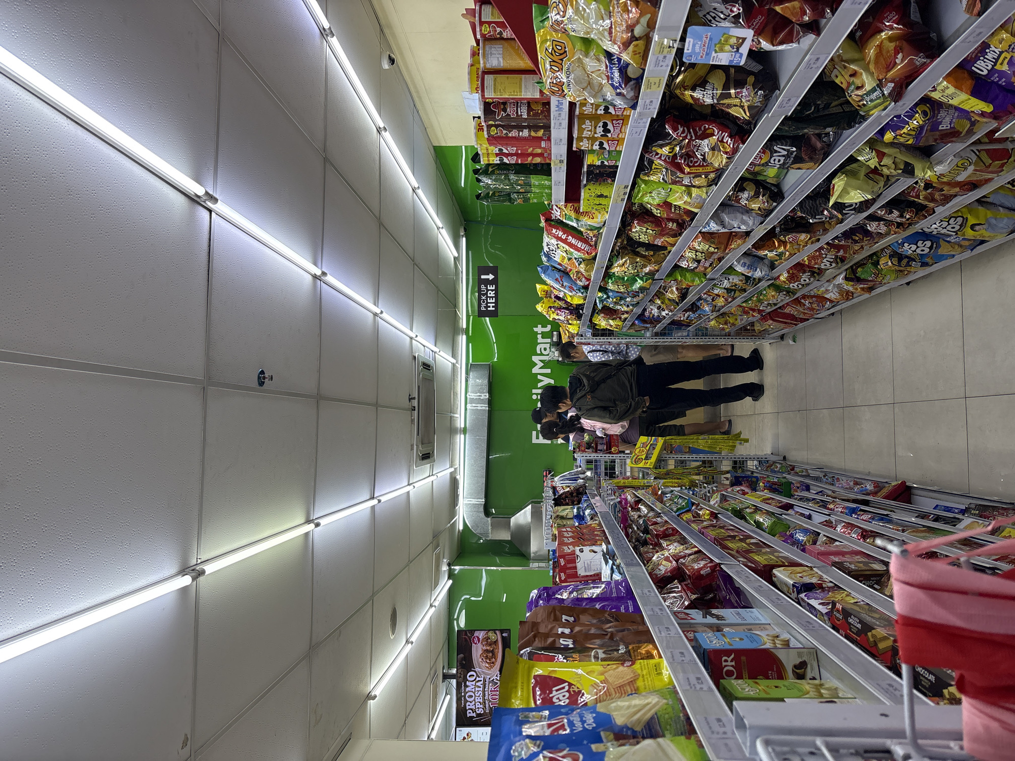 The image depicts the interior of a FamilyMart convenience store located in North Jakarta, Indonesia. The store is well-lit with long fluorescent lights extending across the ceiling. Two aisles of merchandise are prominently featured, with shelves packed with various packaged snacks, drinks, and other convenience items. The aisle on the left displays bags of chips and boxes of cookies, while the aisle on the right is filled with a wider assortment of snacks, including potato chips, crackers, and other packaged food items.
In the middle aisle, three individuals are visible. Two individuals, a male and a female, are walking away from the camera, looking at the merchandise. Another individual, a male, is walking towards the camera. The background of the store features a bright green wall with the "FamilyMart" logo, as well as a "PICK UP HERE" sign with an arrow pointing left. There are also some large industrial ventilation units visible on the green wall. The floor is tiled. The overall impression is that of a typical, well-stocked convenience store.