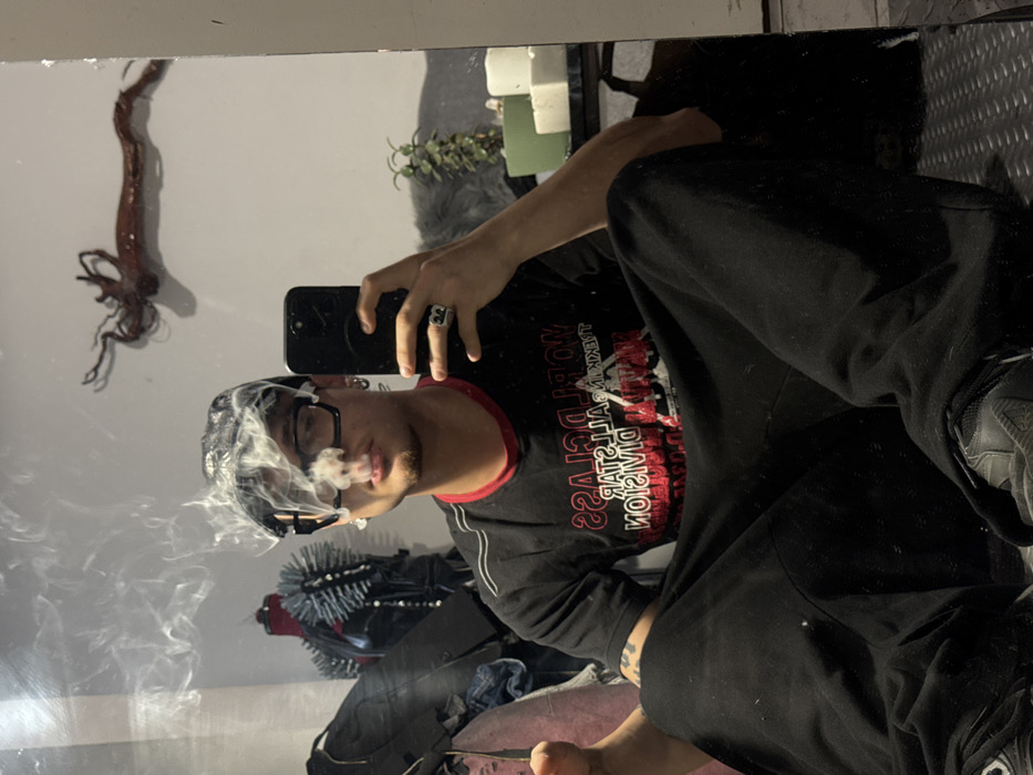 The image is a mirror selfie taken indoors in Sofia, Bulgaria, depicting an individual seated cross-legged on a dark, textured floor covering. The subject, identified as male, is exhaling a cloud of white smoke from their mouth. They wear a black bandana, dark-framed glasses, a black t-shirt with mirrored red and white text "WORLD CLASS STAR DIVISION," black baggy pants, and dark athletic shoes. Multiple earrings are visible on the subject's left ear, and a ring with the letter "B" is worn on the right ring finger, which holds a dark-colored smartphone used to capture the image. Tattoos are visible on the subject's left forearm. The room's background, reflected in the mirror, includes a light-colored wall with a mounted, gnarled, dark branch-like decoration. To the left of the subject, a black spiked garment is displayed on a mannequin, alongside a dark bag. To the right, a small potted plant and two white square objects (possibly candles) are visible on a dark surface.