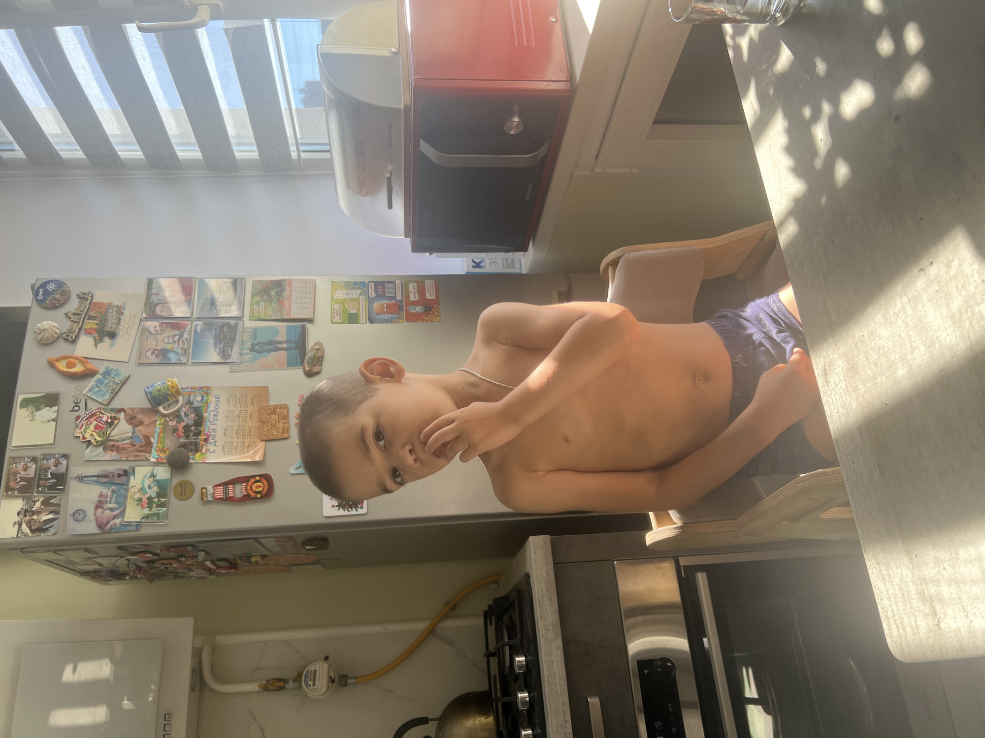 A young boy, likely under 10, is the sole subject in this indoor photograph, taken in a kitchen. He is shirtless and wearing blue patterned shorts. The boy is seated on a wooden chair, leaning forward with his right hand near his mouth, giving the impression he might be eating or thinking. His expression is neutral, and his gaze is directed slightly off-camera. Sunlight streams into the room from the left, casting strong shadows on the wall and floor, indicating it's likely daytime.
The background reveals a kitchen environment. A refrigerator, adorned with numerous magnets and photos, is visible on the left. On top of the refrigerator, a red and silver microwave or retro appliance is placed. To the right of the boy, a countertop is visible, also with shadows from an unseen light source or window. Below the countertop, the top of a stainless steel oven or similar appliance is seen. A gas stove with knobs is also present in the lower part of the frame, along with a yellow gas pipe. The overall setting suggests a domestic kitchen in Volgograd, Russia.
The image is oriented vertically, and the boy's head is towards the top of the frame. The lighting creates a strong contrast between illuminated areas and shadows. There is no discernible text that is clear enough to read within the image.