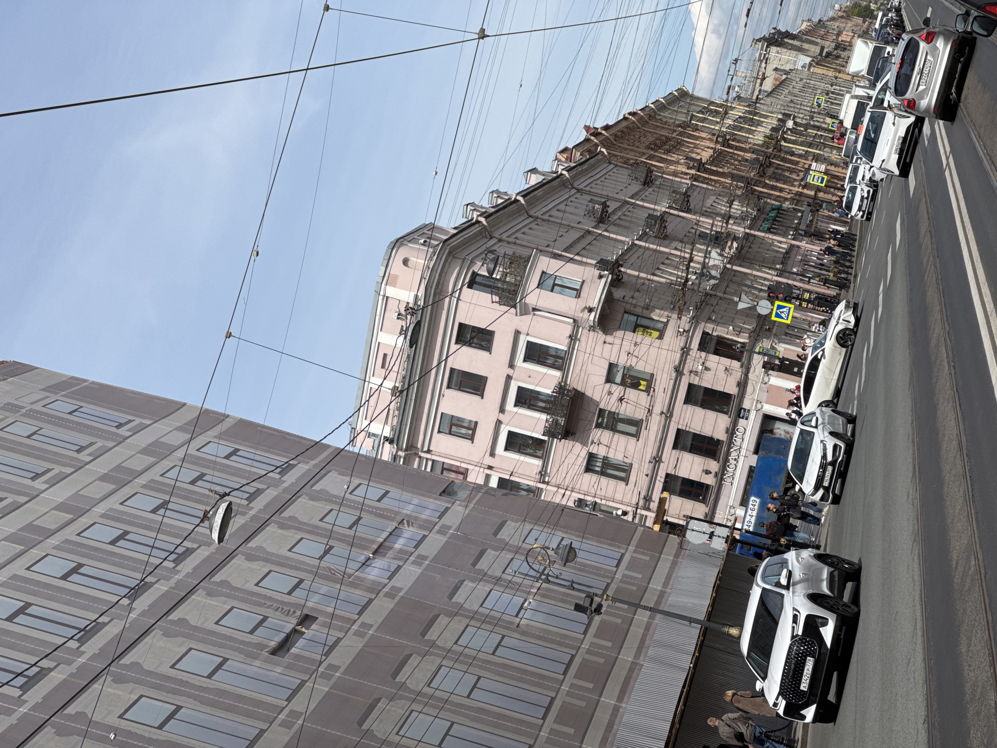 The photograph depicts a street scene in Saint Petersburg, Russia. Buildings of various heights line the street. In the foreground is a modern building with a patterned facade. Behind it is an older, light pink building with multiple windows. Overhead, numerous electrical wires crisscross the sky.
Cars are parked or driving along the street on the right side of the image. People are visible on the sidewalks. A street sign is present near the center of the scene. The sky is partly cloudy, with a blue expanse visible.