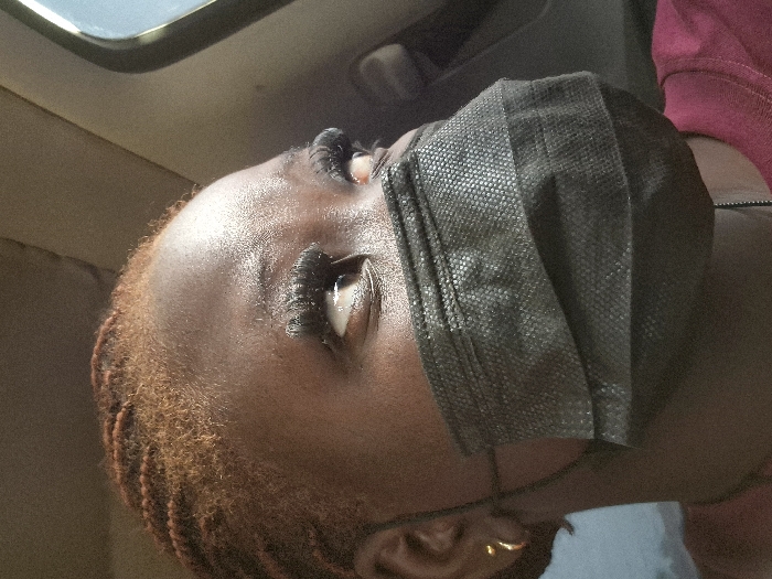 A low-angle shot features a Black person wearing a black face mask. The mask covers the nose and mouth, obscuring the lower half of the face. Dark eyelashes are visible above the mask, and the skin appears smooth. The person has reddish-brown hair. One gold earring is visible. The background suggests an interior space, possibly a car, with a window and a beige seat.