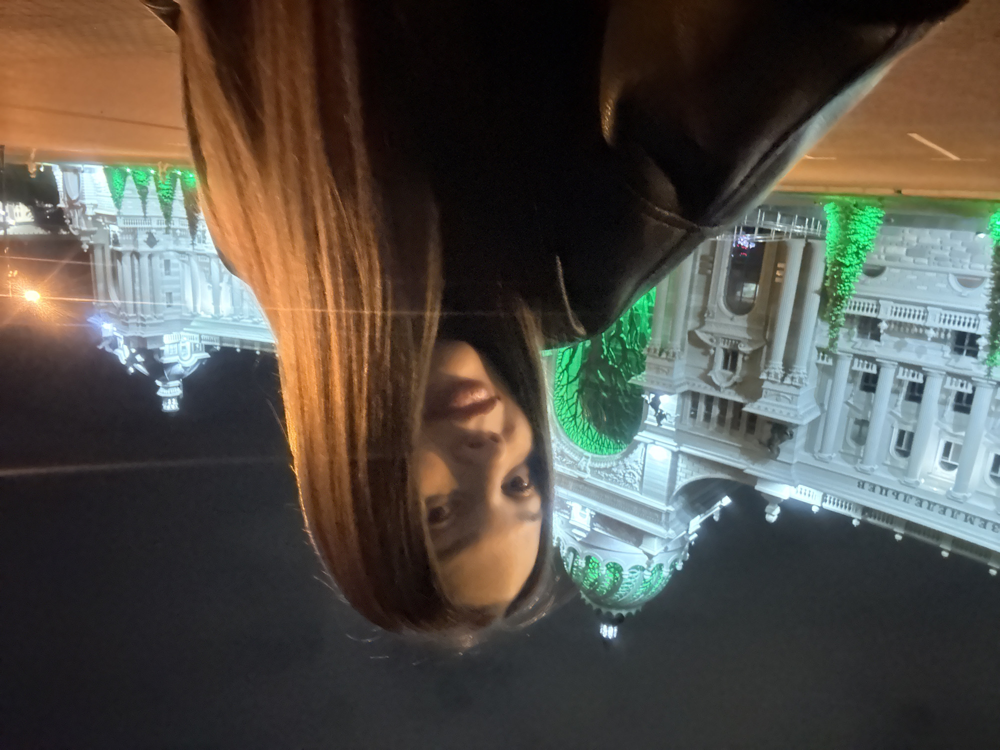 This is a nighttime photograph featuring a young adult woman in the foreground and the Palace of Farmers (Dvoretz Zemledeltsev) in Kazan, Russia, in the background.
The woman occupies the lower right and center of the frame, captured in a close-up, slightly upward-angled shot, suggesting a selfie or a photo taken at arm's length. She has long, straight, light brown hair with lighter highlights, parted in the middle, cascading over her shoulders. Her face is illuminated, showing subtle makeup, and she has a neutral to slightly smiling expression. She is wearing a dark, possibly black, long-sleeved top or jacket.
Behind the woman, the Palace of Farmers is prominently displayed, illuminated against a dark night sky. The building exhibits a grand, ornate architectural style with classical elements such as columns, balustrades, and intricate detailing. The main body of the palace is illuminated with bright white light. The distinctive central archway and the large dome above it are bathed in a vibrant green light, highlighting their decorative features. The text "ЗЕМЛЕДЕЛЬЦЕВ" (Zemledeletsev), which translates to "Farmers," is visible on the left wing of the building. Along the ground in front of the palace, several green, conical lights resembling stylized evergreen trees are arranged.
An asphalt road with white lane markings is visible in the mid-ground, separating the viewer/subject from the palace. In the far distance on the right, streetlights cast a warm glow. The scene depicts a well-lit urban environment at night, emphasizing the architectural grandeur of the Palace of Farmers with its distinctive green illumination.