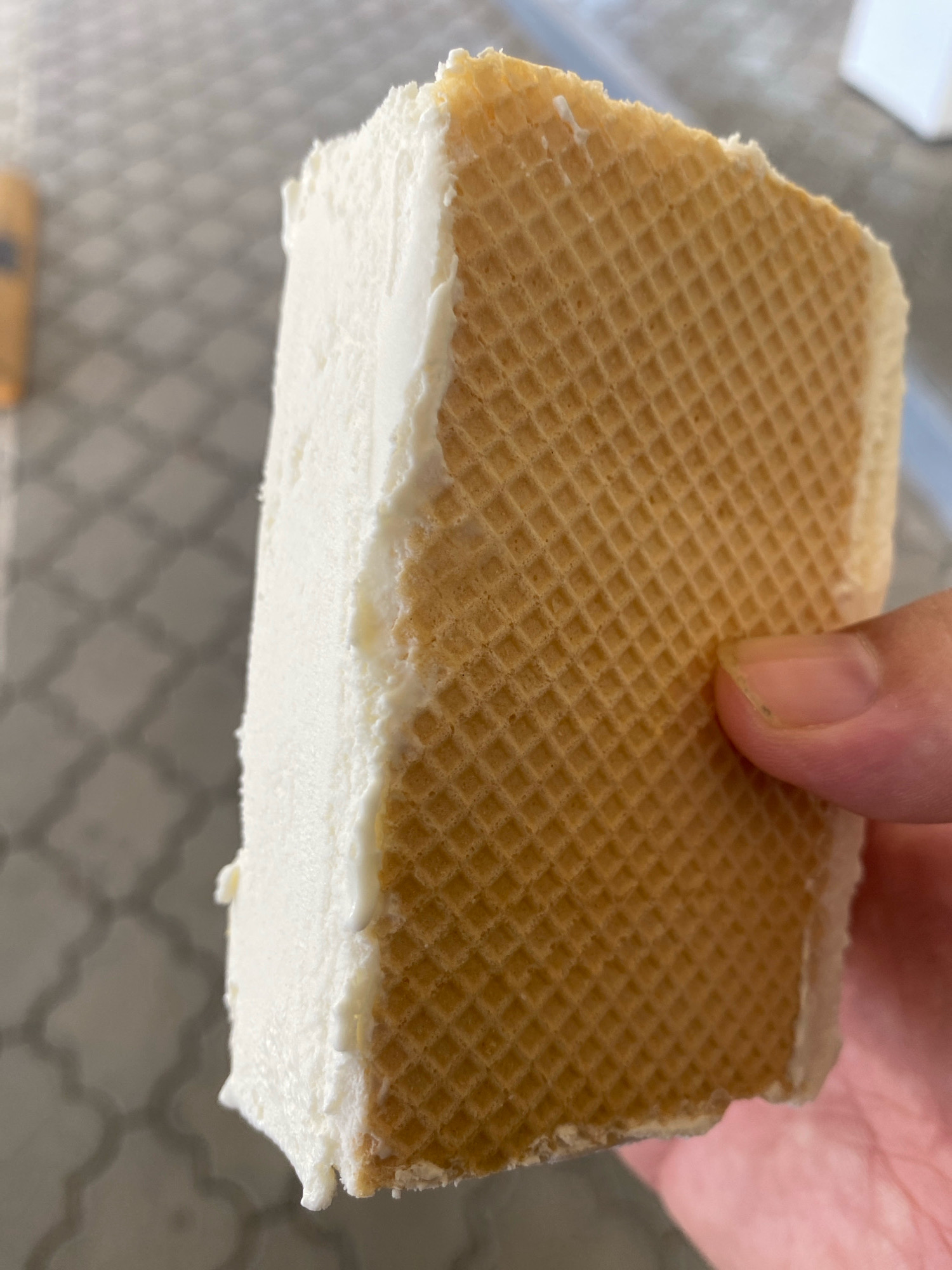 A close-up, eye-level shot shows a hand holding a rectangular ice cream sandwich. The sandwich has a thick layer of white ice cream filling sandwiched between two waffle wafers. The left side of the sandwich, which is not visible, is likely also wafer. The waffle wafers are golden brown and have a grid pattern. The ice cream is slightly softened and has a few drips on the side. The background is a blurred, grey, textured surface, possibly a table with a diamond pattern. There are no identifiable landmarks or features that would allow for specific location within Chișinău, Moldova to be determined.