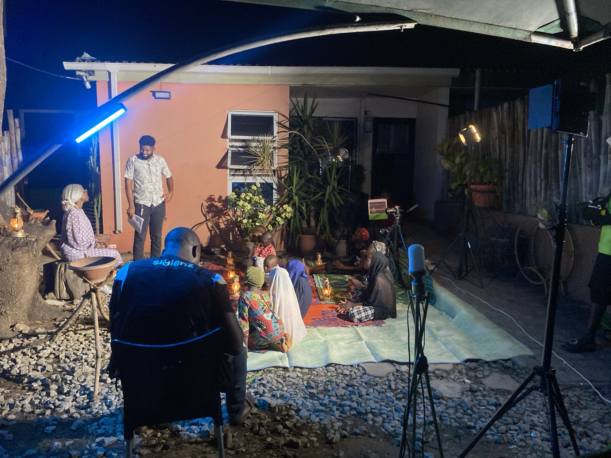 The image depicts a film set, possibly in Barden Yana, Nigeria, at night. A group of people, including women and children dressed in traditional clothing and headscarves, are seated on a patterned rug placed on a light-colored tarp outdoors. Lanterns provide some light, and camera and lighting equipment surround the scene. A man in a patterned shirt and dark pants stands with papers, likely a script, nearby. Another man in a black shirt with white lettering is seated facing the group with his back to the camera. The background includes a building with a window, plants in pots, and a partial view of a fence. The ground is covered in gravel.