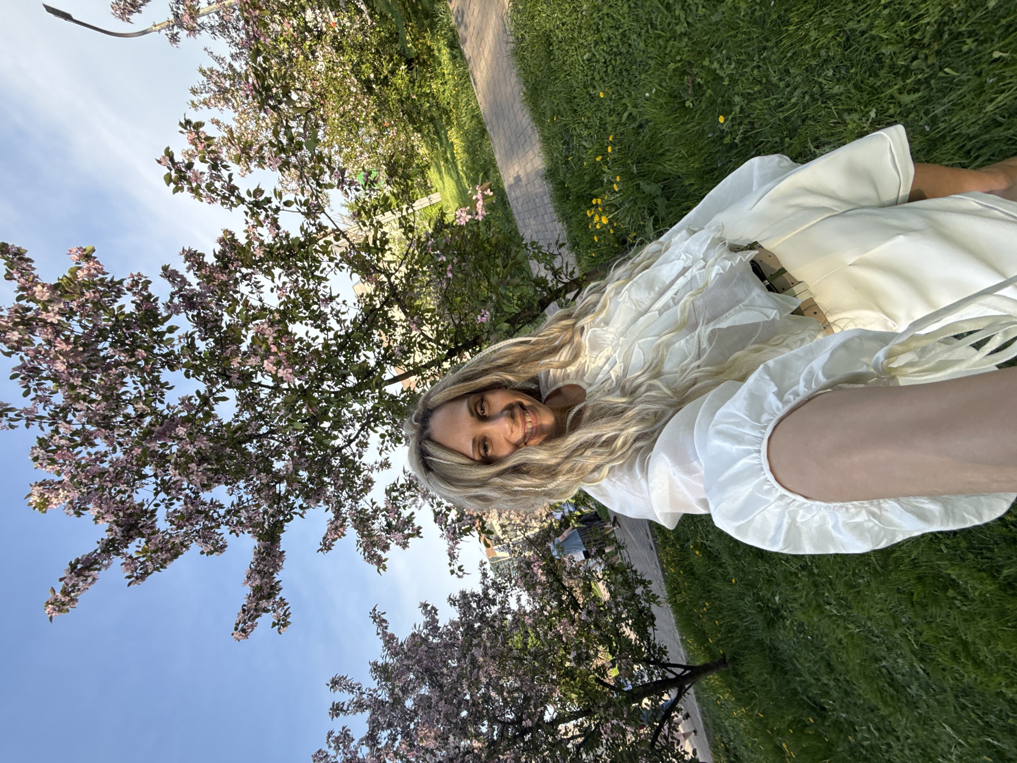 A selfie shows a blonde woman in a white dress lying on grass, with flowering trees visible in the background. The background includes glimpses of a brick walkway, green grass, and what appears to be an urban environment.