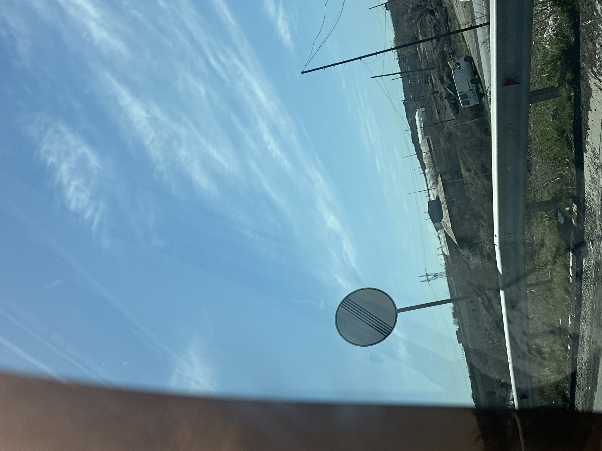 The image shows a view from inside a vehicle looking out the window at a roadside scene. In the foreground, there is a road sign indicating the end of a speed limit zone. Beyond the road sign, there is a road with a guardrail. Further back, there is vegetation and what appears to be a hill or embankment. Power lines and utility poles are visible in the distance. A white van is parked on the side of the road. The sky is blue with streaks of white clouds. The location appears to be a rural or suburban area.