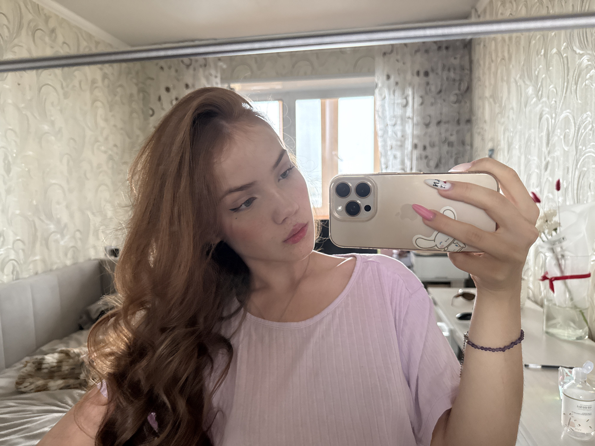 The image depicts a mirror selfie taken by a young female indoors in Naberezhnye Chelny, Russia. The female, appearing in her late teens or early twenties, has long, wavy, reddish-brown hair, parted on her right side, cascading over her shoulders. She is wearing a light lavender, ribbed, short-sleeved top with a round neckline and a delicate silver-colored necklace. Her right hand holds a gold/champagne-colored iPhone, equipped with a clear case that features a white rabbit cartoon sticker on the back. Her visible fingernails are long and oval-shaped; the ring, middle, and pinky fingers are painted bright pink, while the index finger has white polish with a black letter "U" and a small red heart. A bracelet composed of small purple beads is worn on her left wrist. Her face is turned slightly to her left, looking towards the phone screen, and she exhibits winged eyeliner and what appears to be a pink lip tint or gloss.
The reflected environment is a residential room. The walls are covered with light-colored wallpaper featuring an ornate, textured scrollwork or damask pattern. A multi-pane window with white, patterned curtains is visible in the background, allowing bright daylight into the room. To the left, a bed with a light-colored, padded headboard (featuring horizontal stitching) and light grey satin-like bedding is present, along with a crumpled, multi-toned patterned blanket or throw. To the right, a white flat surface, possibly a table or shelf, holds various items: a clear glass vase or jar containing white flowers or decorative elements and tied with a red ribbon, a small clear plastic bottle with a white pump dispenser (possibly hand sanitizer), a dark pen or stylus, and partially visible sunglasses with dark lenses. A horizontal silver-colored bar or rail is visible across the top portion of the mirror's reflection.