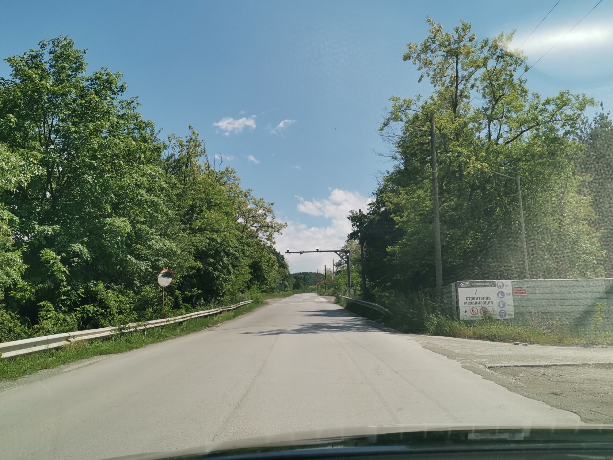 The image captures a first-person perspective from a vehicle, looking down a two-lane asphalt road in Momin Prohod, Bulgaria, under a bright blue sky with scattered white clouds. Lush green deciduous trees line both sides of the road, forming a natural tunnel. The road surface shows visible wear, cracks, and patches.
On the left side of the road, a white metal guardrail is present, and further along, a circular convex mirror, likely for blind spot visibility, is mounted on a pole. In the distance, a dark metal gantry-like structure spans across the road, fitted with what appear to be lights or sensors, potentially indicating a checkpoint or monitoring station.
To the right, a utility pole stands, and behind it, a large white sign is clearly visible. The sign, which appears to be a construction safety board, features the text "GP СТРОИТЕЛНА КОМПАНИЯ" (GP Construction Company) at the top left. The main text below it reads "СТРОИТЕЛНА МЕХАНИЗАЦИЯ" (Construction Mechanization), surrounded by several warning and mandatory pictograms. These include symbols for "no entry", "no smoking", "hard hat required", "eye protection", "ear protection", "safety vest", "safety shoes", and "gloves", among others. The clear, bright daylight suggests it is midday or early afternoon. No people are visible in the tranquil, natural setting, which is punctuated by signs of human infrastructure and construction activity.