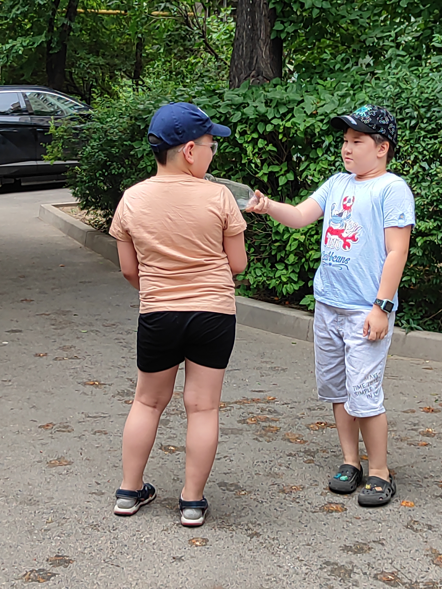 Two young boys are standing on a paved surface in what appears to be a residential courtyard or park area in Almaty, Kazakhstan. The boy on the left, facing away from the camera, wears a blue baseball cap, glasses, a peach-colored t-shirt, and black shorts. The boy on the right, facing the other boy, wears a patterned baseball cap, a light blue t-shirt with a graphic print, and light gray shorts. He is holding a clear object, possibly a bottle or container, and reaching out towards the other boy, suggesting an interaction or exchange. The background consists of dense green foliage and trees. A portion of a dark-colored car is visible on the left side of the image. The ground is paved with asphalt and scattered with fallen leaves. The overall scene depicts a casual outdoor moment between two children.