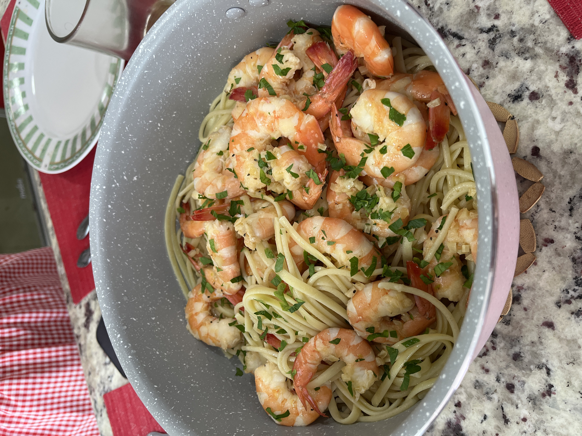 This close-up image showcases a large, grey speckled pot filled with a generous serving of shrimp scampi or garlic butter shrimp and linguine pasta. The plump, pinkish-orange shrimp are interspersed throughout the light-colored pasta, all glistening with what appears to be a buttery garlic sauce. Freshly chopped green parsley is sprinkled abundantly over the dish, providing a vibrant contrast.
The pot has a light pink or rose gold colored exterior rim visible on the right side. The dish is placed on a speckled countertop, likely granite or quartz, featuring a mix of white, grey, and reddish-brown tones. In the upper left corner, a white plate with a green striped pattern around its edge is partially visible, alongside a clear glass or cup. A red and white checkered cloth or placemat can be seen on the bottom left. Additionally, a few dried, brown, claw-like objects are visible on the countertop behind the pot on the right, possibly decorative items.
The scene appears to be set in a home, possibly a kitchen or dining area, illuminated by bright, even lighting, suggesting daytime. The meal looks freshly prepared and appetizing, evoking a sense of warmth and domestic comfort. No visible text is present in the image. Although the context provided indicates the location is Barrio de Nuevo León, Mexico, there are no specific visual cues within the photograph itself to confirm this geographical detail.