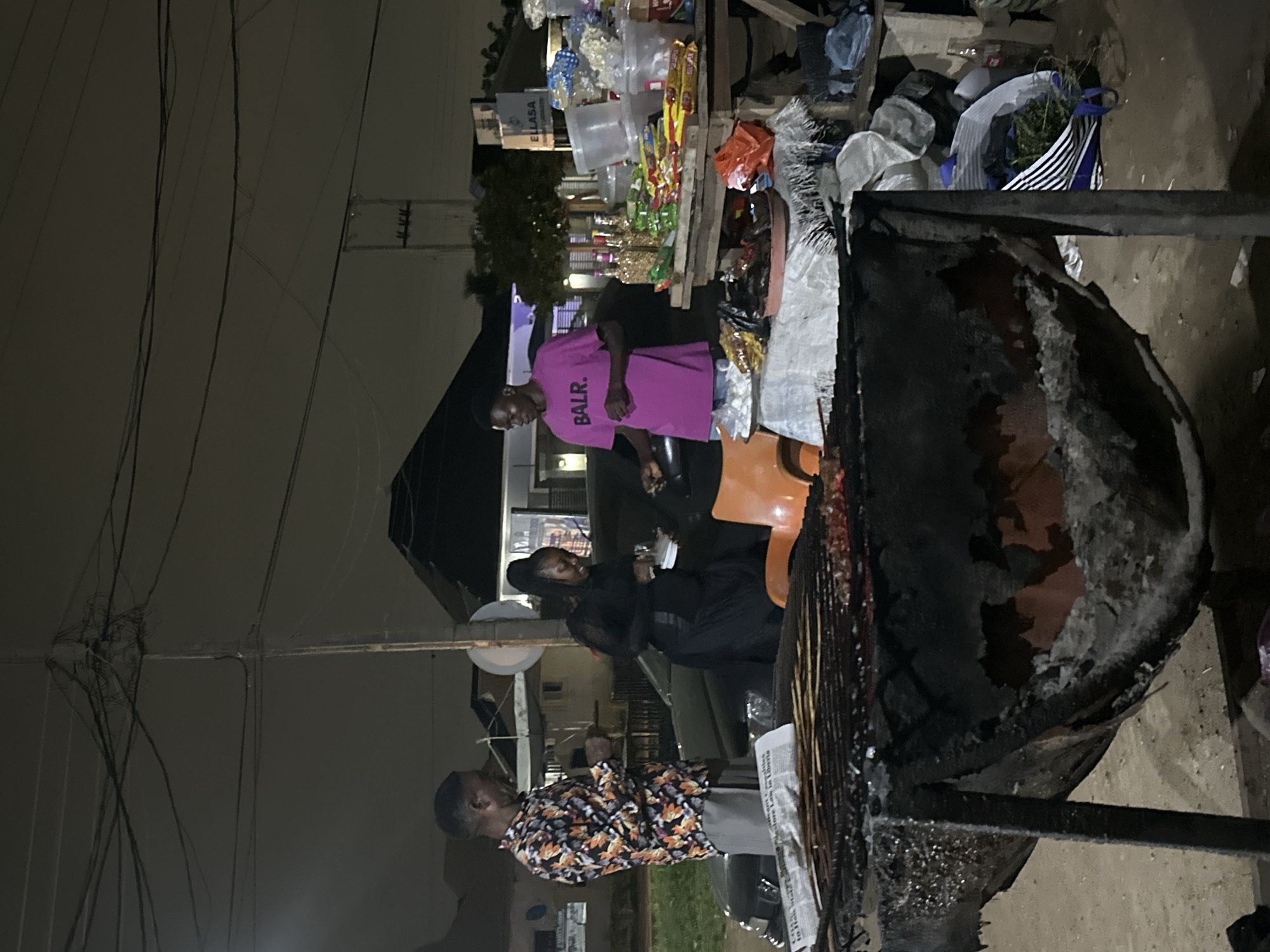 The image depicts a nighttime scene on a street in Abuja, Nigeria, possibly near a roadside stall. In the foreground, a large, rustic grill is visible, filled with what appears to be meat skewers cooking over embers.
Three individuals are the central focus. On the left, a man wearing a vibrantly patterned long-sleeved shirt and grey trousers stands facing right, holding a phone. To his right, a woman in a long black dress is engaged in a transaction, holding some items in her hands. On the far right, a young man in a bright pink t-shirt with the logo "BALR." printed on it stands near a wooden table laden with various goods, including packaged snacks, bottles, and plastic containers. He appears to be interacting with the woman.
The environment suggests a bustling street setting with overhead power lines creating a dense network against the dark sky. In the background, buildings and trees are dimly visible, hinting at the urban landscape of Abuja. The lighting is artificial, primarily from streetlights and the glow of the grill, casting long shadows and creating a somewhat dramatic ambiance. The overall impression is of a typical evening street food vending operation in Nigeria's capital.