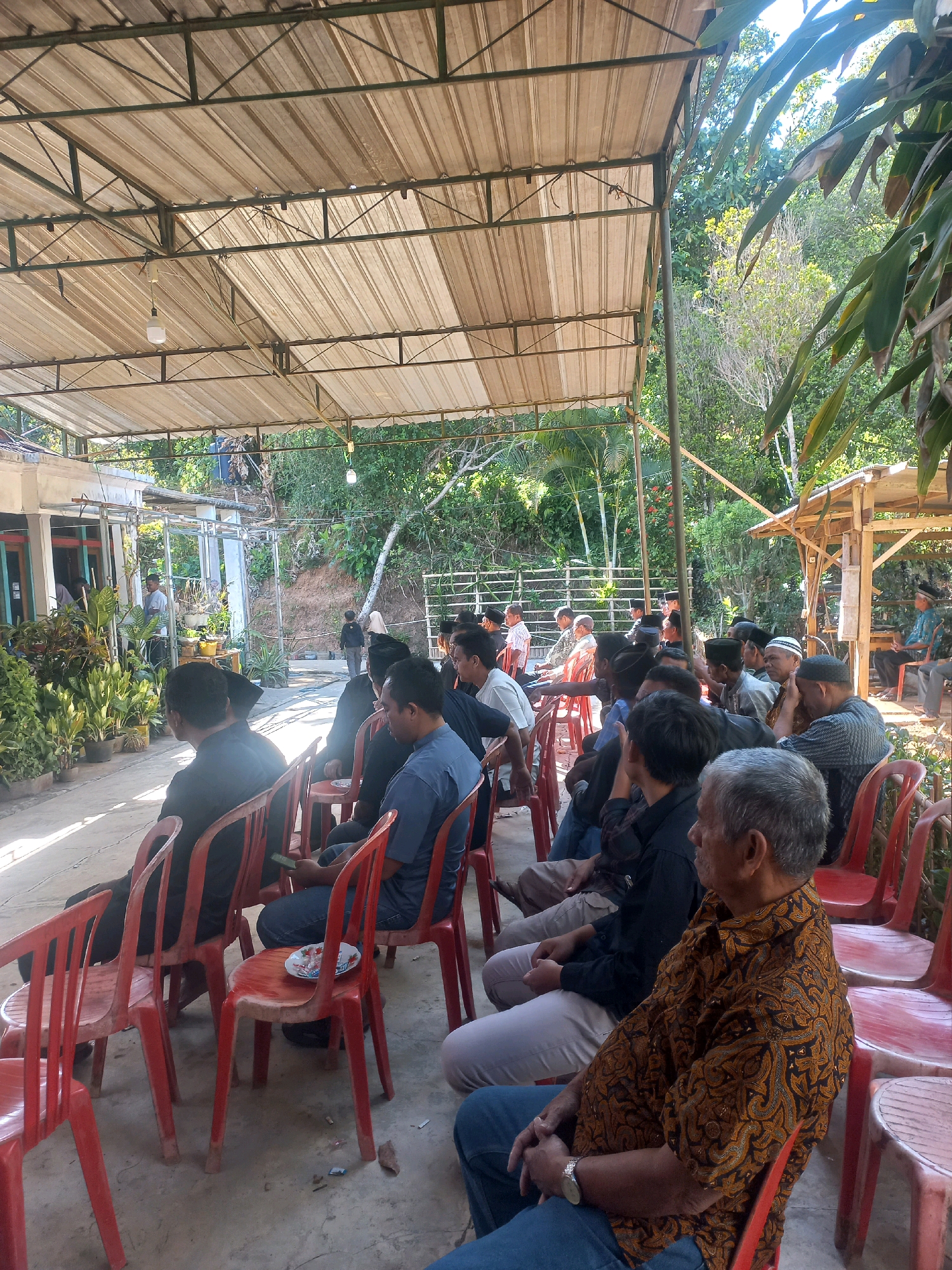 The image depicts an outdoor gathering of men seated on red plastic chairs under a large, open-sided canopy supported by a metal frame. The canopy is constructed of corrugated metal roofing. The setting appears to be a community or religious event. The ground is paved with concrete. In the background, there are lush green trees and vegetation, suggesting a rural or semi-rural environment. To the left, a building with a white facade and glass doors is visible, surrounded by potted plants. To the right, a small wooden structure, possibly a gazebo or a shelter, is present. Several men are visible, mostly in the foreground and midground, dressed in casual attire, with some wearing traditional Islamic caps (peci). They are facing forward, seemingly listening to a speaker or observing an event. The overall atmosphere is one of communal assembly. The mention of Sukoharjo, Indonesia, indicates the geographical context of this scene, which is likely a local village or town square where such events are held.