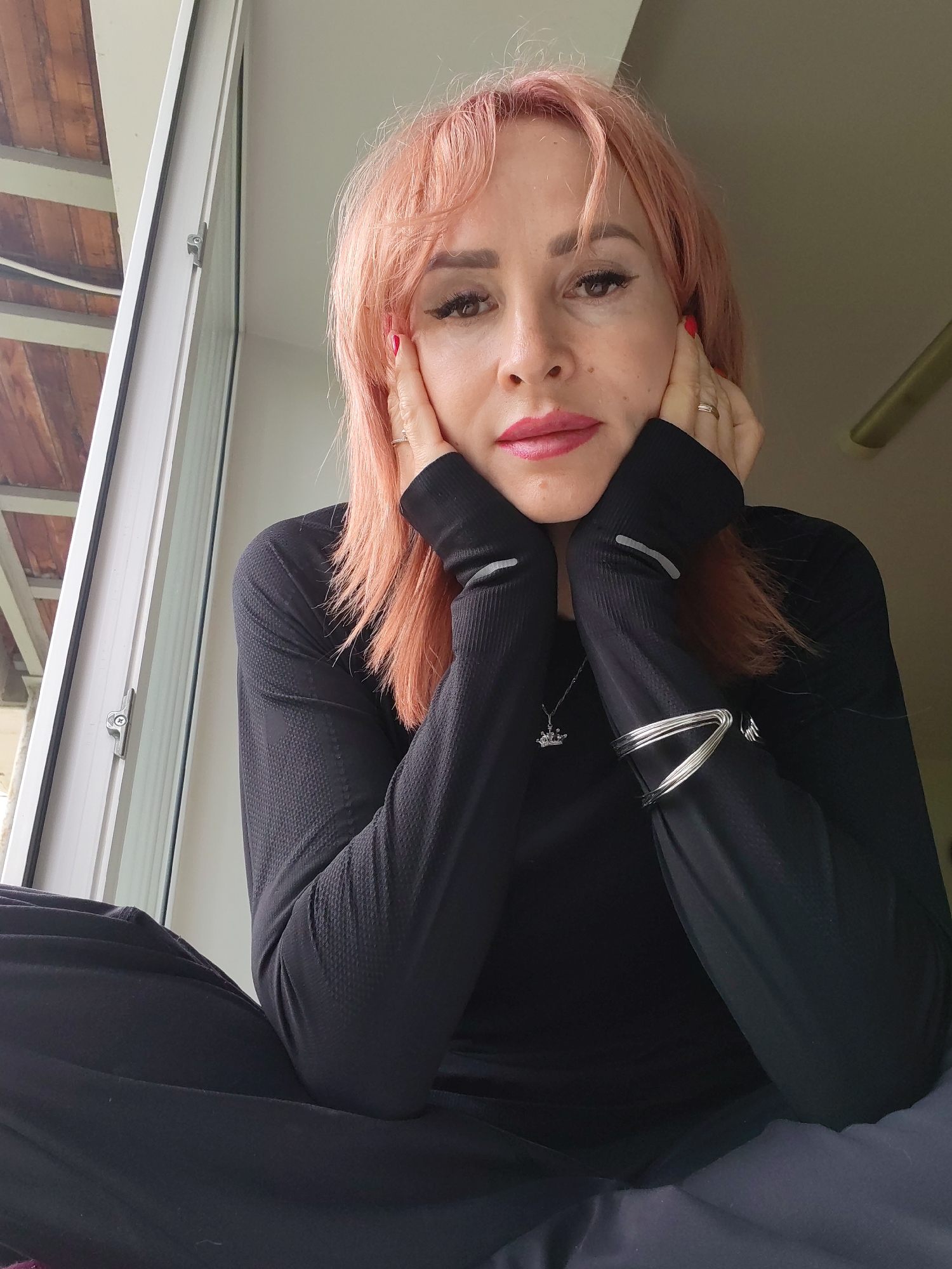 A woman with pink hair is sitting in front of a window in what appears to be a modern apartment. She is wearing a black long-sleeved shirt and black pants. Her hands are resting on her face, and she is looking at the camera. The room is sparsely furnished, and the only visible details in the environment are a window with blinds and a ceiling fan. The lighting suggests it is daytime. There is no discernible information to suggest the location is in the city of Chaikovsky, Russia.