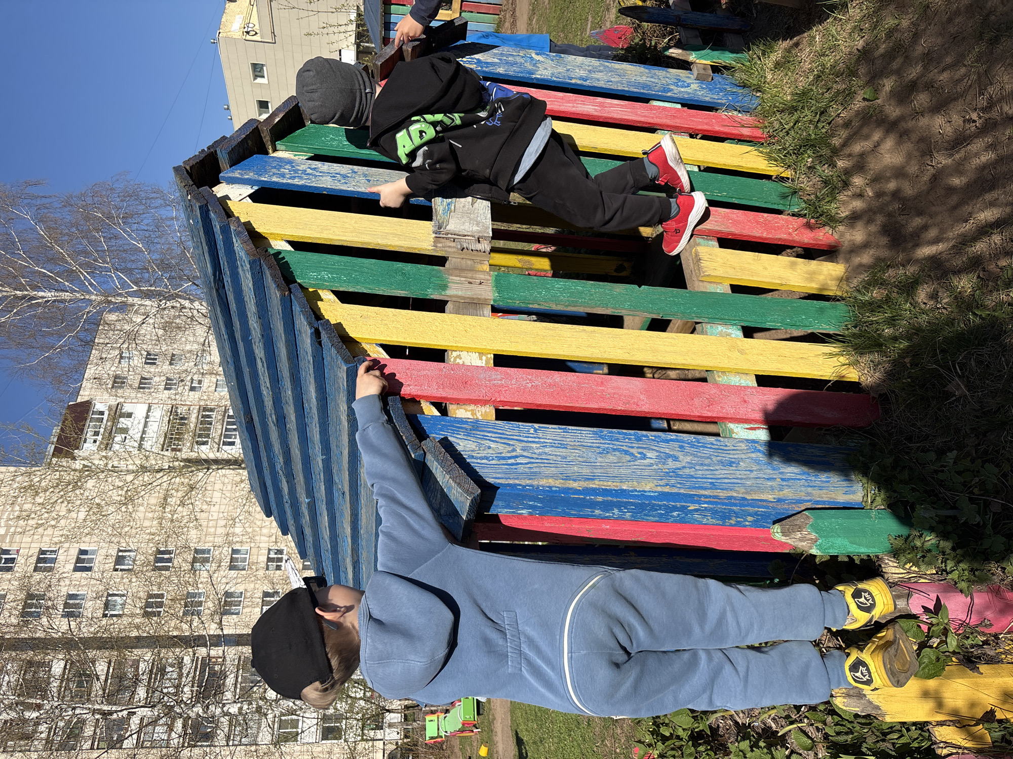 Two children are playing on a brightly colored wooden structure in an outdoor setting in Chaikovsky, Russia. The structure is painted in blue, yellow, green, and red. One child is wearing a blue hoodie and black hat, with yellow shoes, and is reaching upward towards the structure. The other child is wearing a black hoodie with a green design, black pants, a gray hat, and red shoes, and is lying atop the structure, appearing to climb or move across it. In the background is an apartment building with multiple windows. Bare trees are visible in the foreground. The sky is blue. The ground has patches of grass and dirt.