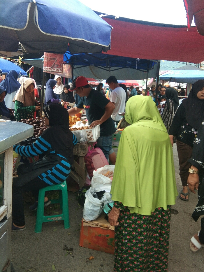 This image captures a lively outdoor market scene in Baleendah, Indonesia, under the shade of various awnings and tarps, primarily blue and red. The setting is bustling, filled with people engaged in commerce.
In the foreground, to the left, a person wearing a black hijab and a blue and black striped long-sleeved top is seated on a bright green plastic stool, attending a stall with dark, round fruits, likely salak. On the right, a woman seen from behind wears a vibrant lime green hijab and a dark green floral-patterned long skirt, standing and observing the market. Her left hand, appearing somewhat aged, is visible. White plastic bags filled with goods and an orange crate are on the ground near her.
In the midground, a man in a red cap and a black t-shirt is actively working at a stall, arranging or serving light-colored fruits, possibly rambutan or longan, from a white plastic basket. To his left, several women, mostly wearing hijabs, are browsing or purchasing items from stalls. One woman with a light brown hijab and green patterned top is reaching for fruit. To the right, more market-goers, including women with children, move through the busy aisles.
The scene depicts a typical market environment with vendors and customers. The lighting suggests it is daytime. A red sign is partially visible under a blue awning, with legible text that appears to read "ODENG KUAH." The overall atmosphere is busy and functional, characteristic of a local market.