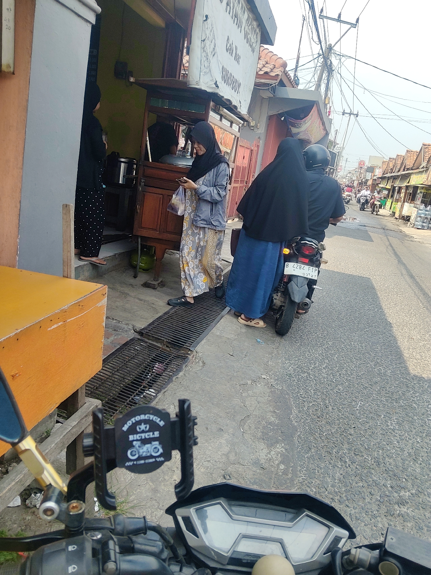 In Mekarmukti, Indonesia, a street scene unfolds with several individuals and vehicles. Two women in headscarves are visible near a food stall. One woman in a black headscarf and polka-dot pants stands to the left. To her right, a woman in a grey jacket and patterned skirt holds a phone and a bag, standing by a wooden food cart. A gas tank is positioned beneath the cart. In the foreground, a motorcycle with a phone mount is parked, its handlebars and dashboard visible. Further down the street, a couple rides a motorcycle, the passenger wearing a black headscarf and blue skirt, and the driver wearing a helmet. The street is lined with shops and stalls, and numerous other motorcycles and people are present, indicating a busy urban environment. Electrical wires crisscross overhead.