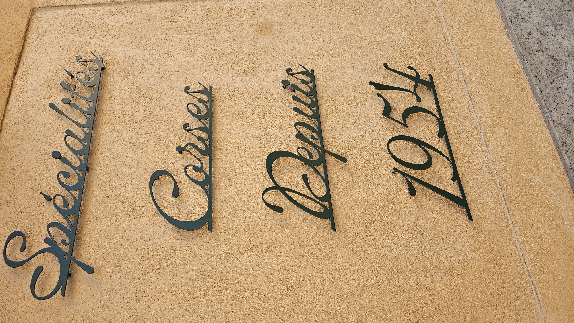 The image shows a sign on a textured, light brown wall. The sign consists of four lines of dark green, cursive text. The top line reads "Spécialités". The second line reads "Corses". The third line reads "Depuis". The fourth line reads "1954". The text is mounted on the wall with small dark green screws. The wall appears to be exterior stucco. Below the wall, there is a concrete or stone surface that is speckled gray. The scene is outdoors, suggesting it is a storefront or building facade. The sign indicates a business that specializes in Corsican products and has been in operation since 1954. This suggests the location is in Corsica, and given the context of L'Île-Rousse, France, it is likely a shop or restaurant there.