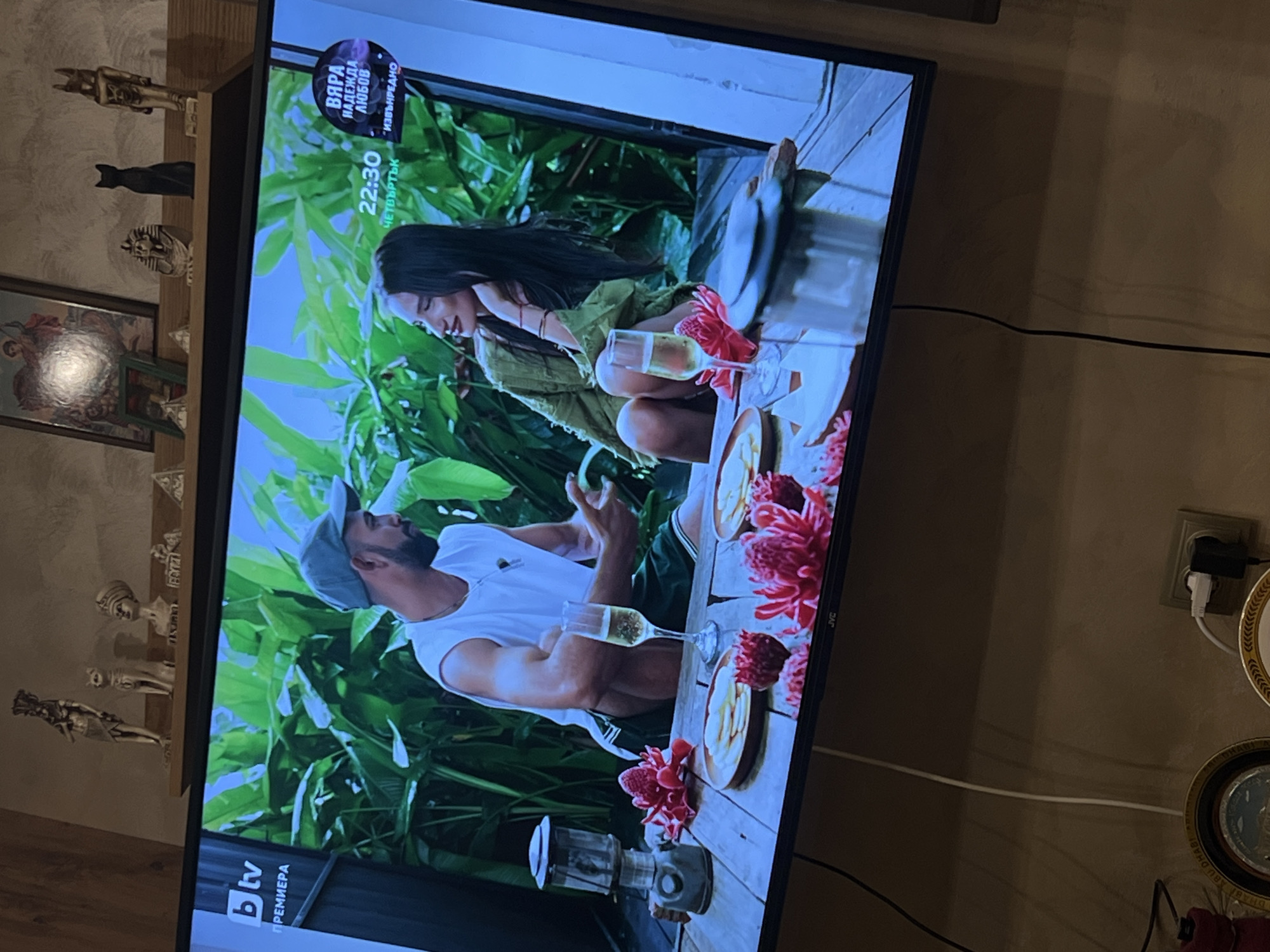 A television displays a scene depicting a man and a woman seated at a table with food and drinks, amidst lush greenery. The TV rests on a wooden surface where decorative figurines are placed. The setting is presumably indoors in Popintsi, Bulgaria. A wall outlet with plugs and decorative plates are also visible in the background.