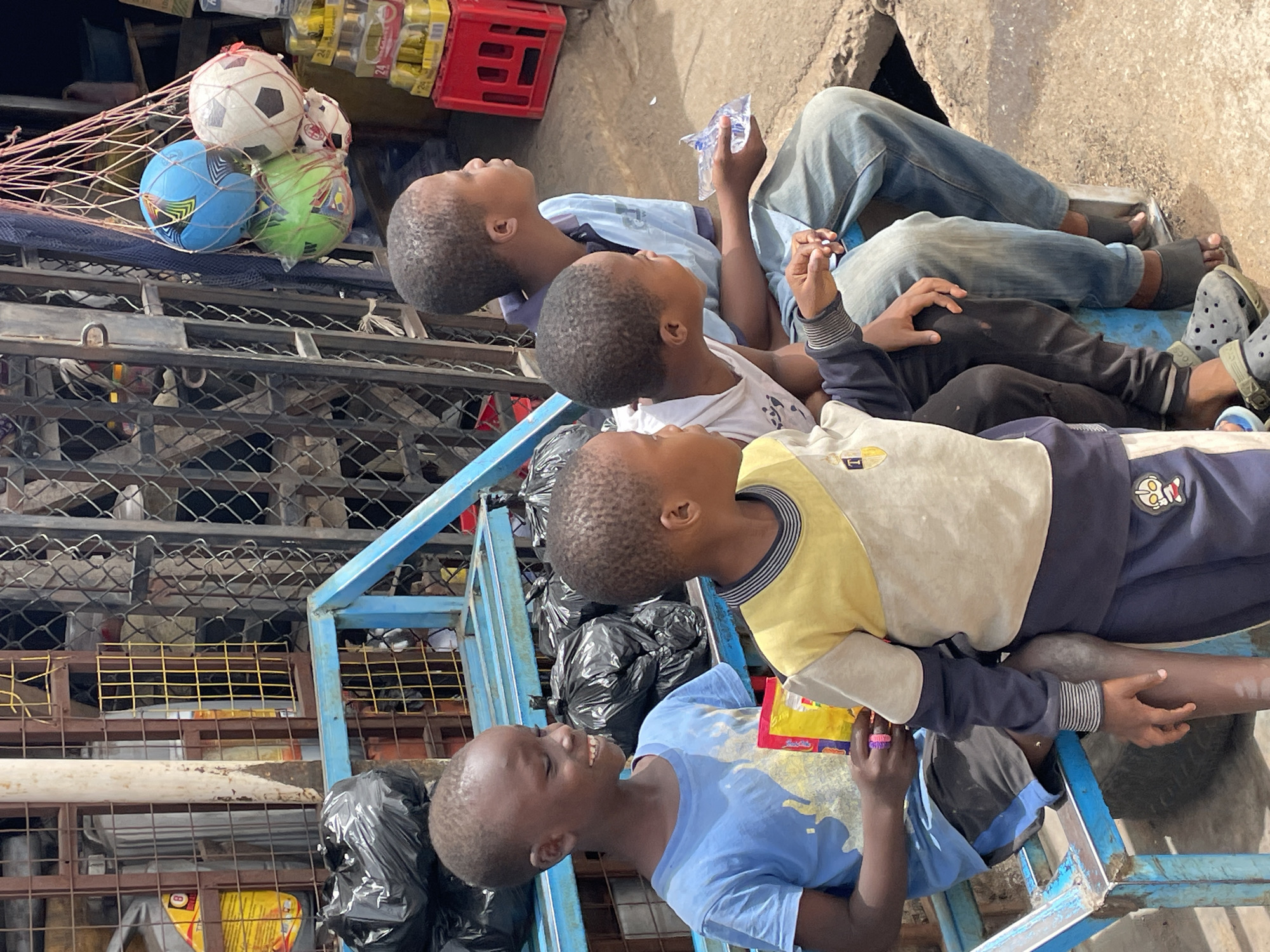 In Jos, Nigeria, four Black children are positioned on a blue metal structure. One child, wearing a light blue shirt, smiles and holds an item in their left hand. Another child, clad in a yellow and navy blue shirt, has their hand resting near the other. A third child wears a white shirt with black patterns, while the fourth is dressed in a light blue shirt and blue jeans. The children are looking upwards. Above the structure, a stack of soccer balls in a net sits near a red crate. Black trash bags are nearby. The background features metal fencing and concrete.